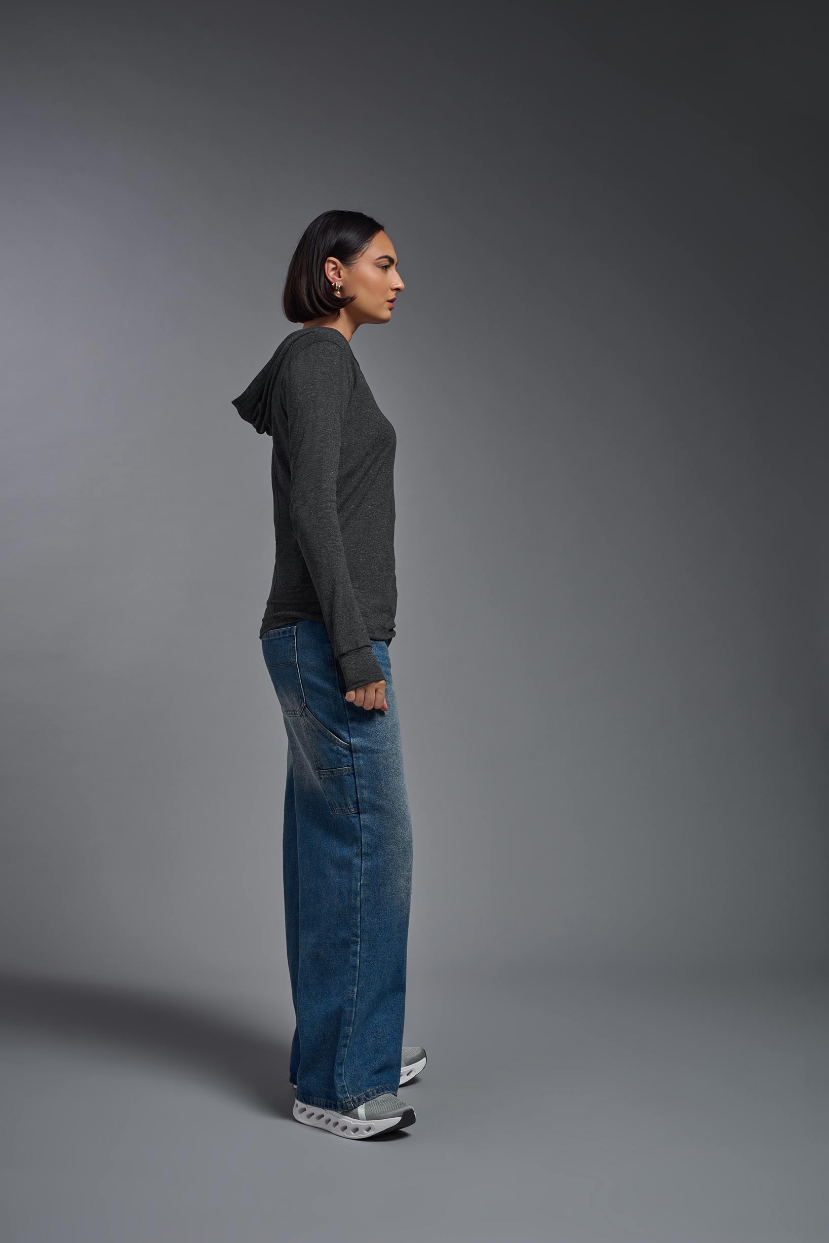 A female model wearing the women's Heather Hoodie Shirt in the color dark heather charcoal, made out of 88% polyester and 22% treated cotton, showing the relaxed fit from a side view. This hoodie shirt is comfortable and made in the USA. #dark-heather-charcoal