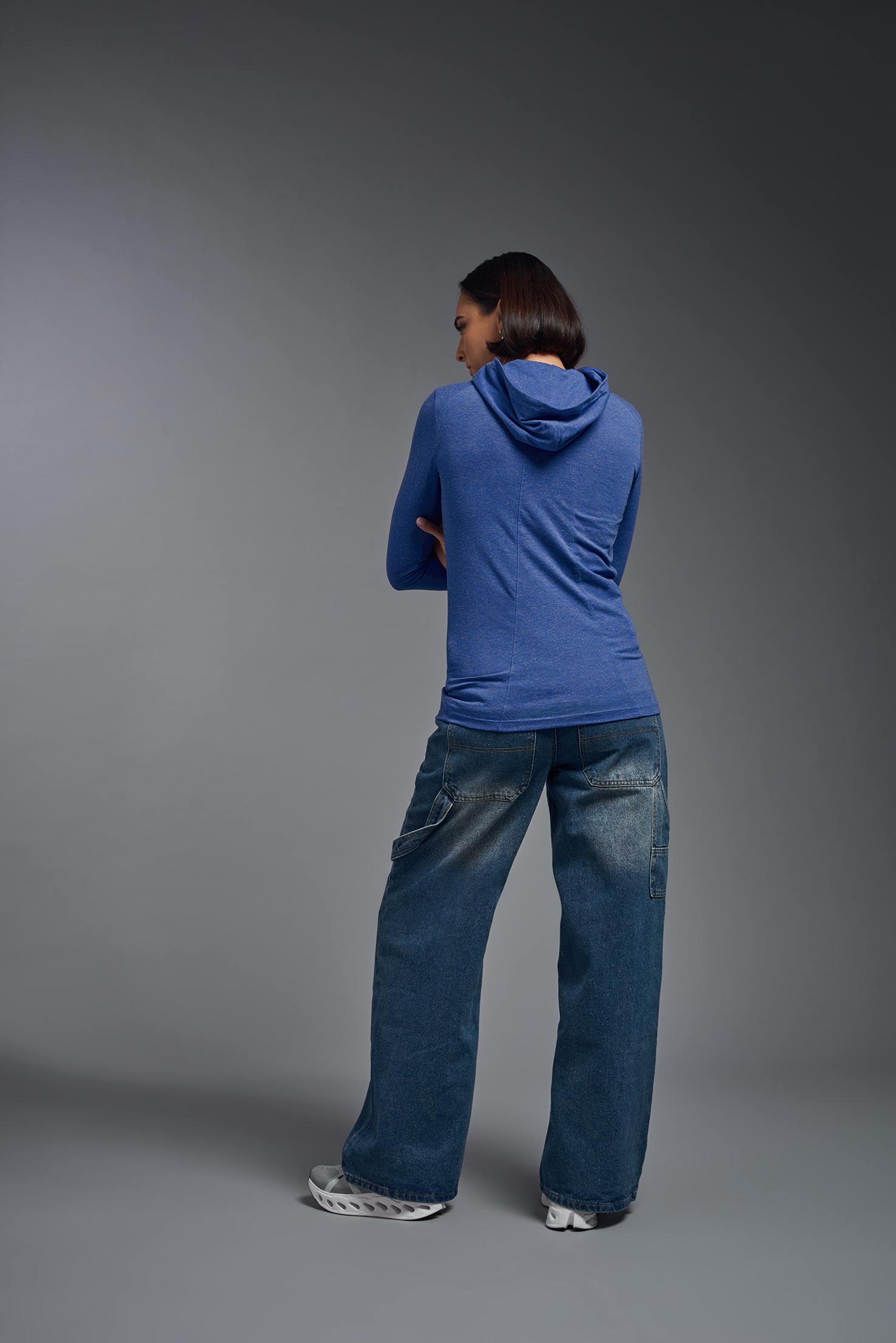A female model wearing the women's Heather Hoodie Shirt in the color dark heather royal blue, made out of 88% polyester and 22% treated cotton, showing the relaxed fit from a back view. This hoodie shirt is comfortable and made in the USA. #dark-heather-royal