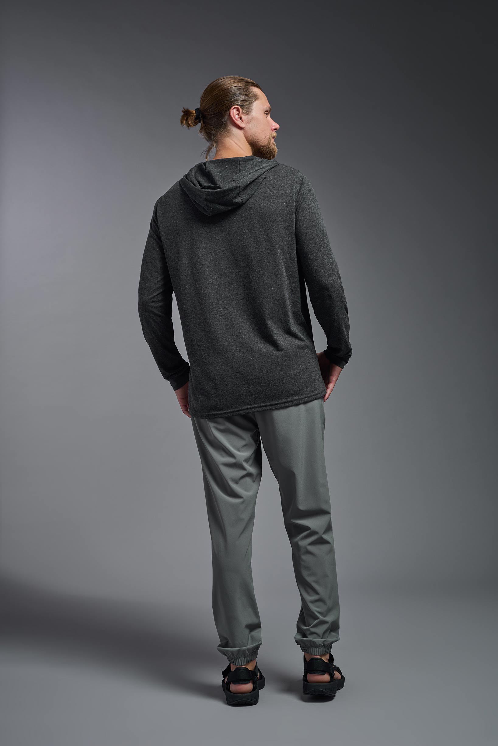 A male model wearing the men's Heather Hoodie Shirt in the color dark heather charcoal, made out of 88% polyester and 22% treated cotton, showing the relaxed fit from a back view. This hoodie shirt is comfortable and made in the USA. #color_dark-heather-charcoal