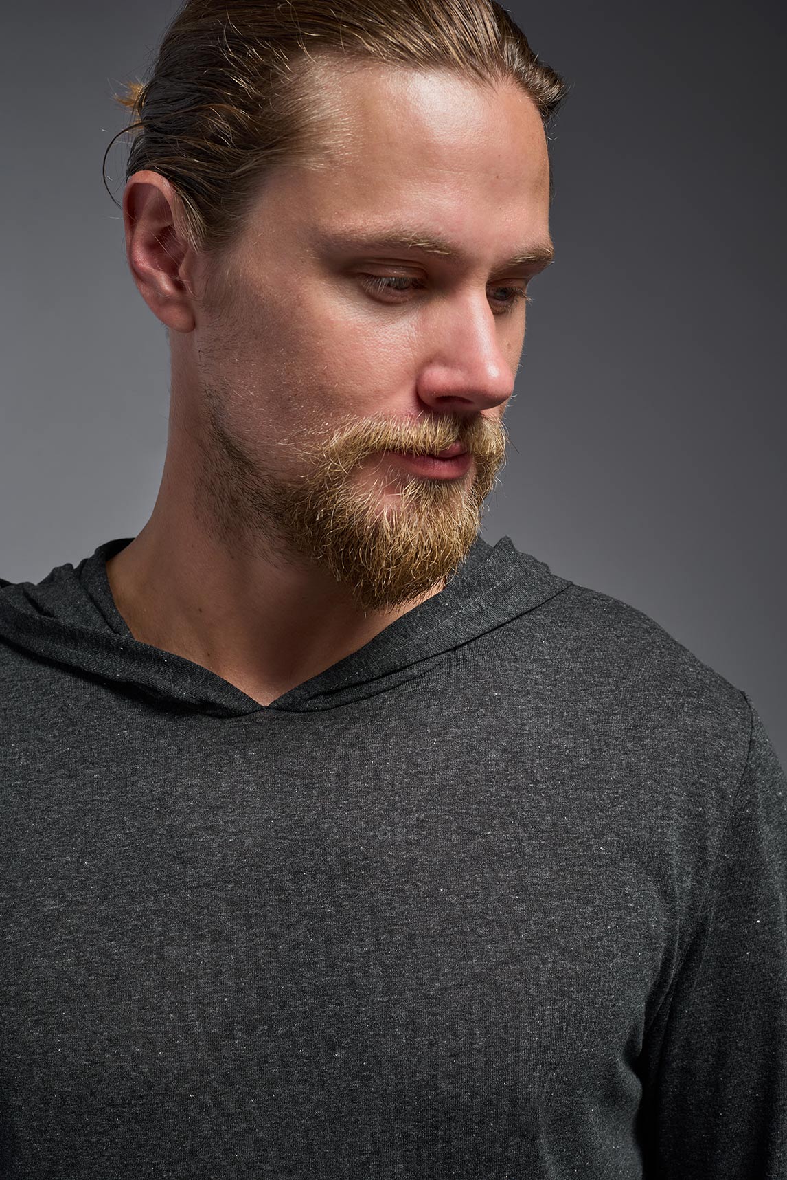 A male model wearing the men's Heather Hoodie Shirt in the color dark heather charcoal, made out of 88% polyester and 22% treated cotton, showing the relaxed fit from a closeup front collar view. This hoodie shirt is comfortable and made in the USA. #color_dark-heather-charcoal
