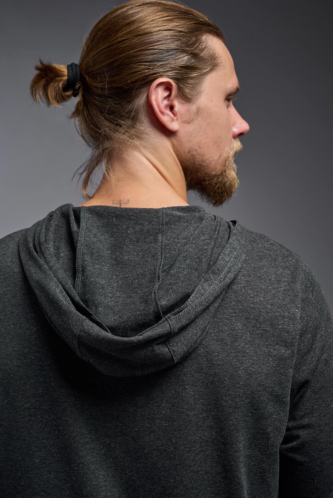 A male model wearing the men's Heather Hoodie Shirt in the color dark heather charcoal, made out of 88% polyester and 22% treated cotton, showing the relaxed fit from a closeup back hoodie view. This hoodie shirt is comfortable and made in the USA. #color_dark-heather-charcoal