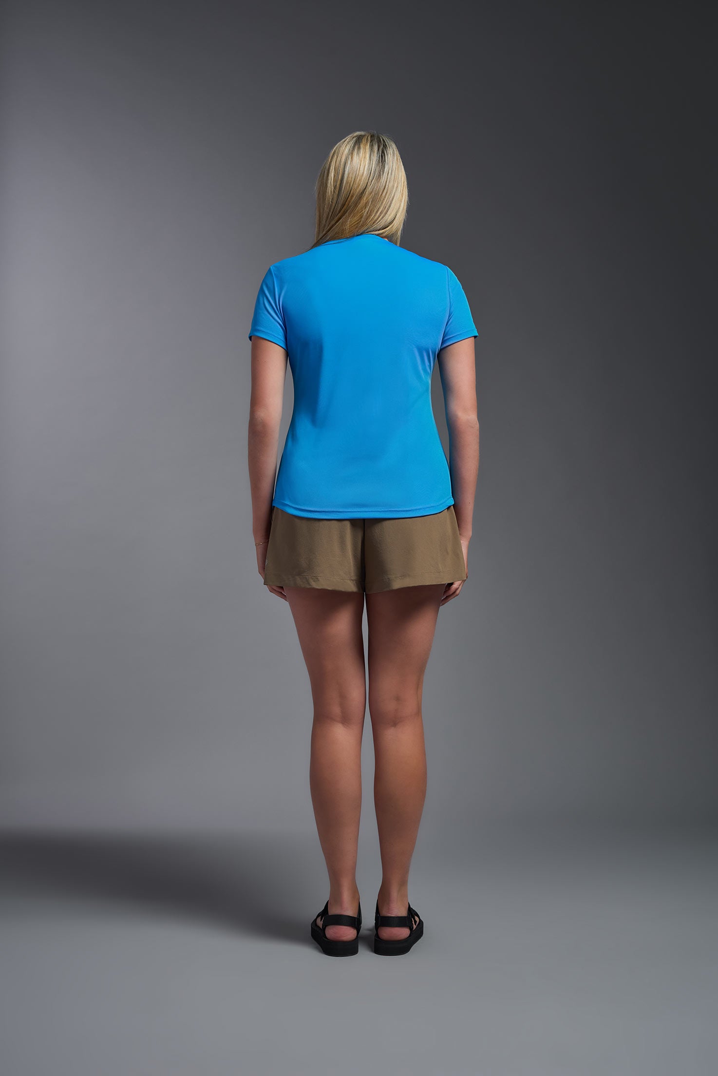 A female model wearing the women's DriMax Crewneck Expert Tee in the color safety blue, made out of 100% polyester, showing the relaxed fit from a back view. This shirt is quick-drying, moisture-wicking, offers up to 30 upf protection and is made in the USA. #safety-blue