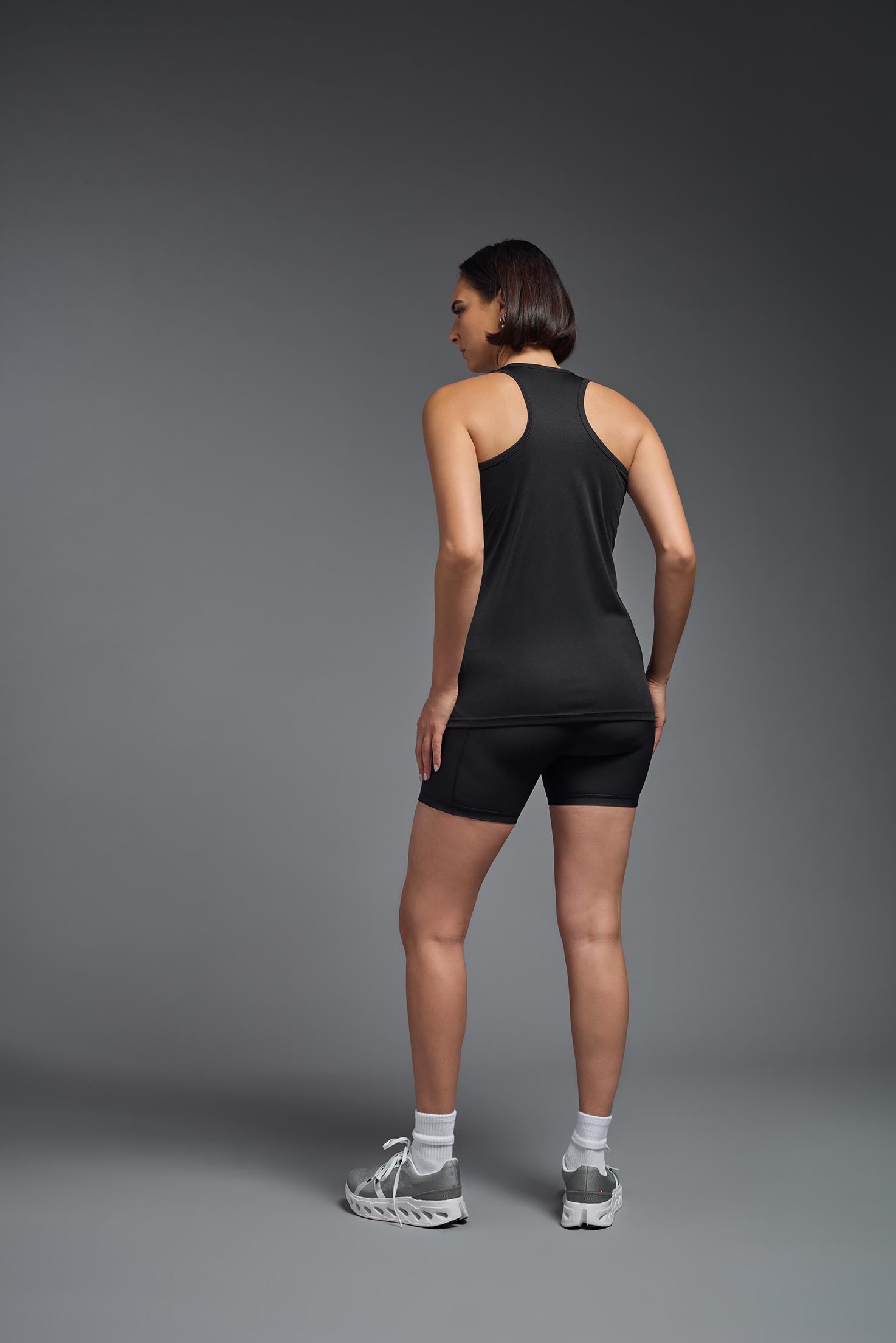 A female model wearing the women's DriMax Endurance Racerback Tank in the color black, made out of 100% polyester, showing the relaxed fit from a back view. This tank is quick-drying, moisture-wicking, with up to 30 UV protection and made in the USA. #black