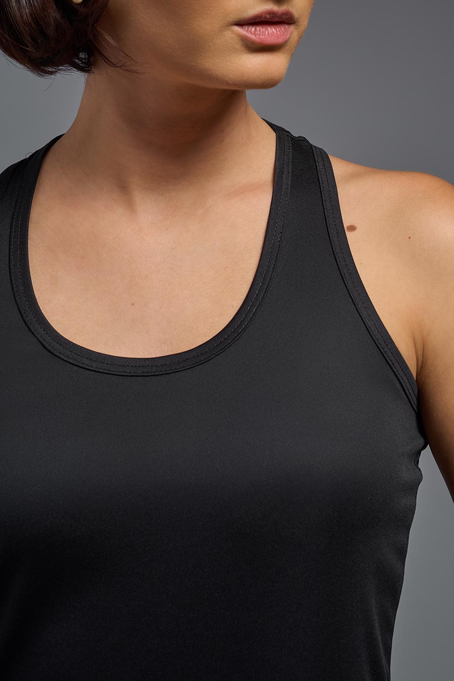 A female model wearing the women's DriMax Endurance Racerback Tank in the color black, made out of 100% polyester, showing the relaxed fit from a close-up collar view. This tank is quick-drying, moisture-wicking, with up to 30 UV protection and made in the USA. #black