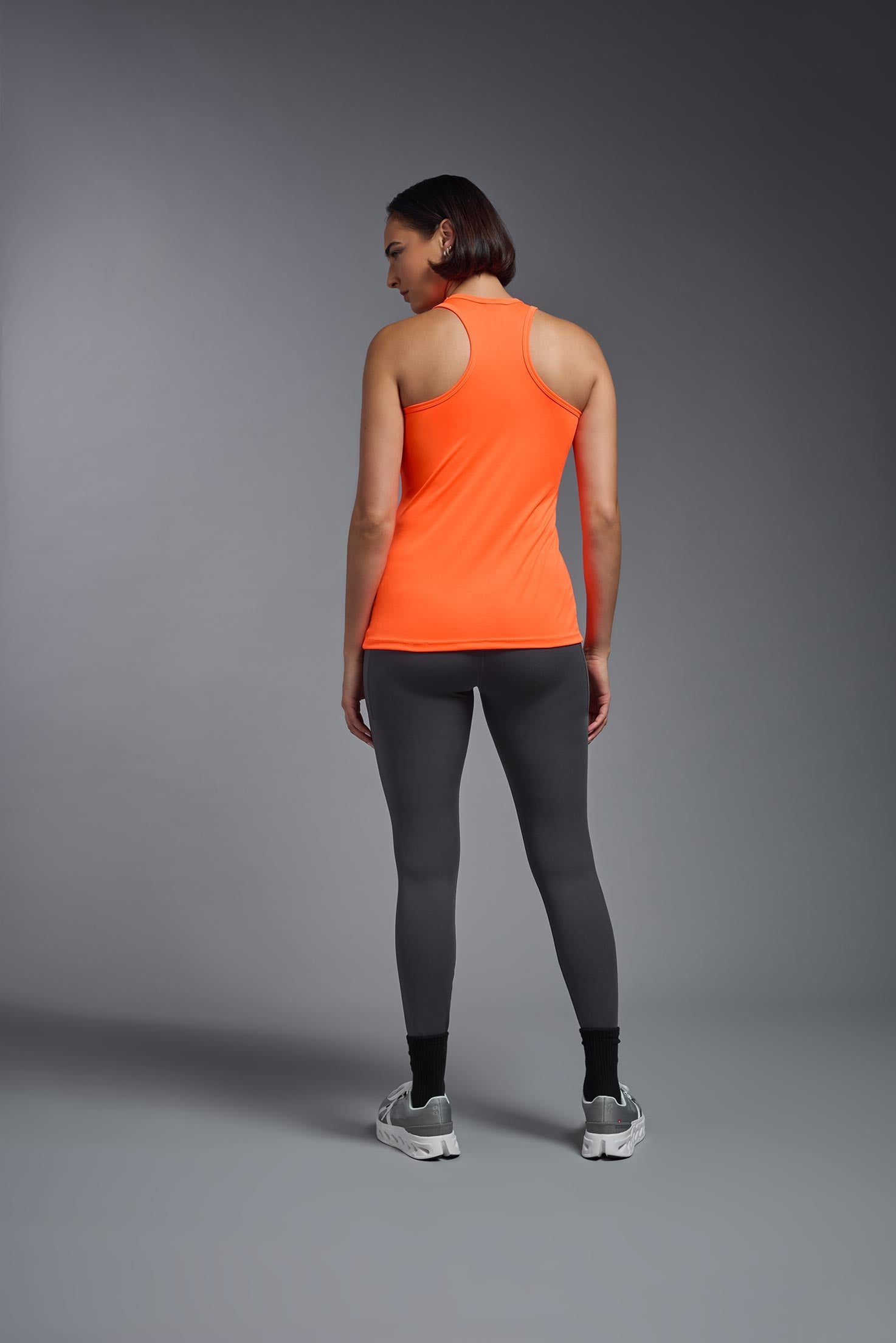 A female model wearing the women's DriMax Endurance Racerback Tank in the color safety orange, made out of 100% polyester, showing the relaxed fit from a back view. This tank is quick-drying, moisture-wicking, with up to 30 UV protection and made in the USA. #safety-orange