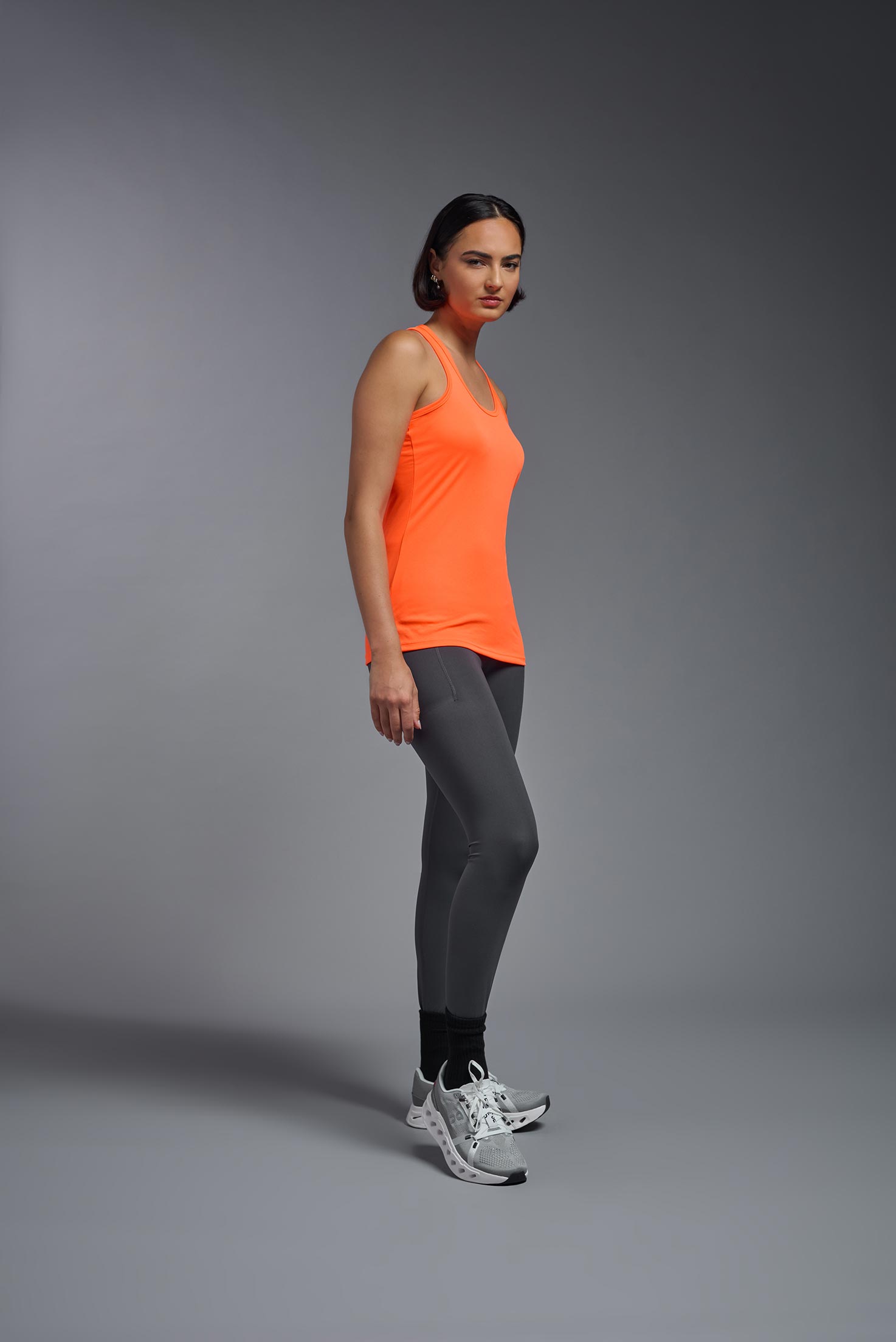 A female model wearing the women's DriMax Endurance Racerback Tank in the color safety orange, made out of 100% polyester, showing the relaxed fit from a side view. This tank is quick-drying, moisture-wicking, with up to 30 UV protection and made in the USA. #safety-orange