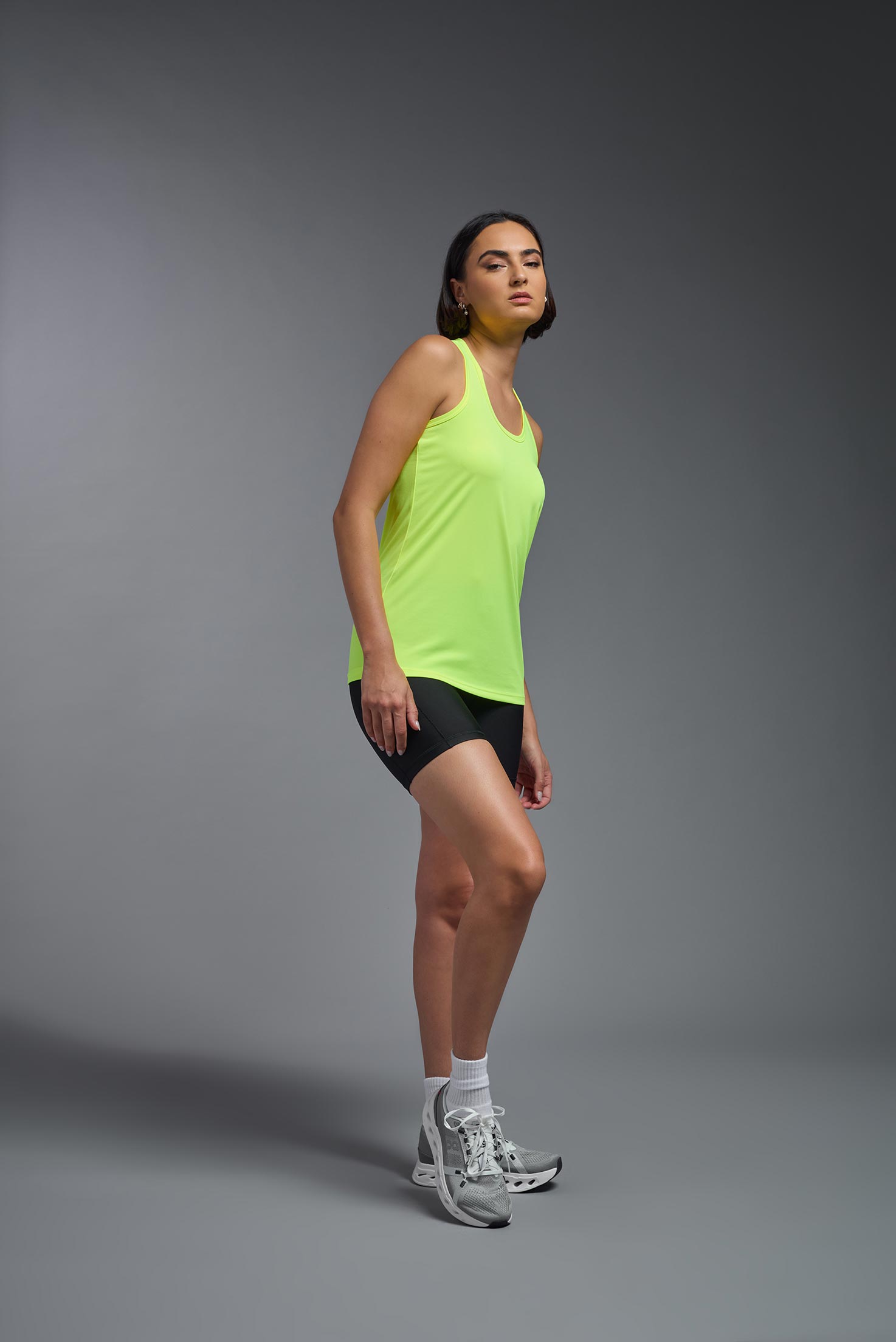 A female model wearing the women's DriMax Endurance Racerback Tank in the color safety yellow, made out of 100% polyester, showing the relaxed fit from a side view. This tank is quick-drying, moisture-wicking, with up to 30 UV protection and made in the USA. #safety-yellow