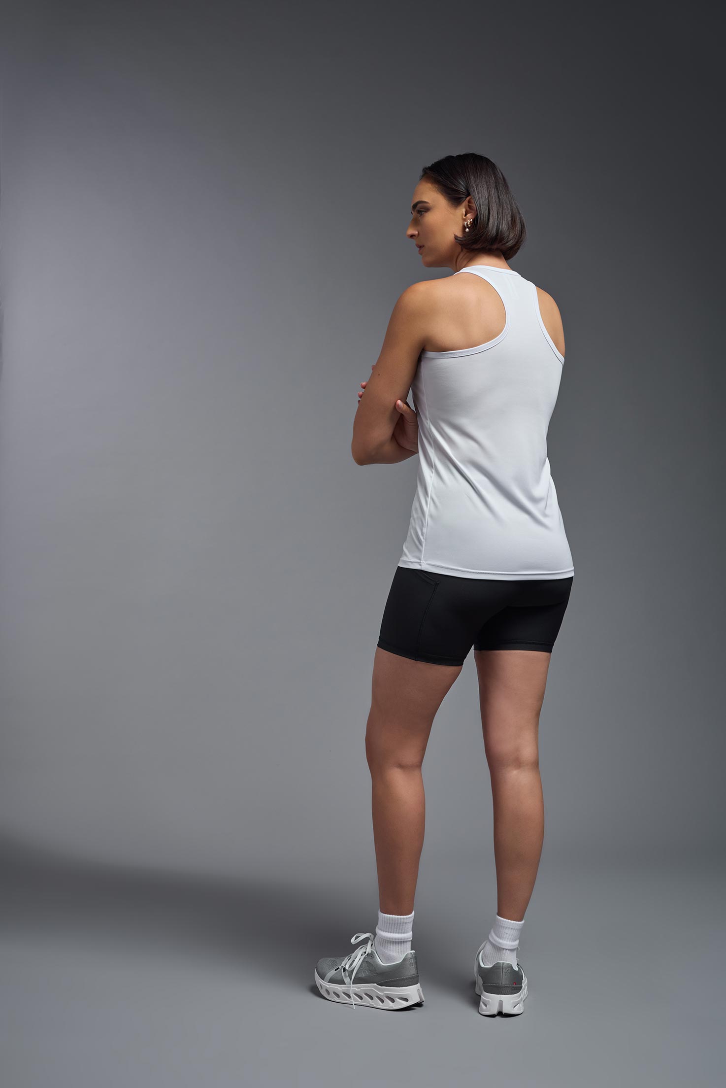 A female model wearing the women's DriMax Endurance Racerback Tank in the color white, made out of 100% polyester, showing the relaxed fit from a back view. This tank is quick-drying, moisture-wicking, with up to 30 UV protection and made in the USA. #white