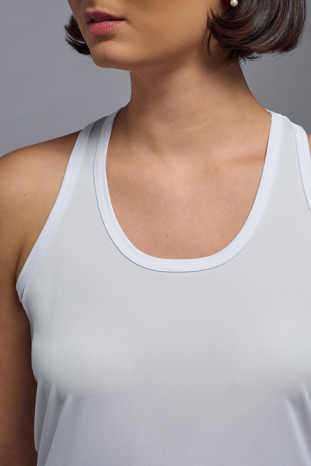 A female model wearing the women's DriMax Endurance Racerback Tank in the color white, made out of 100% polyester, showing the relaxed fit from a close-up collar view. This tank is quick-drying, moisture-wicking, with up to 30 UV protection and made in the USA. #white