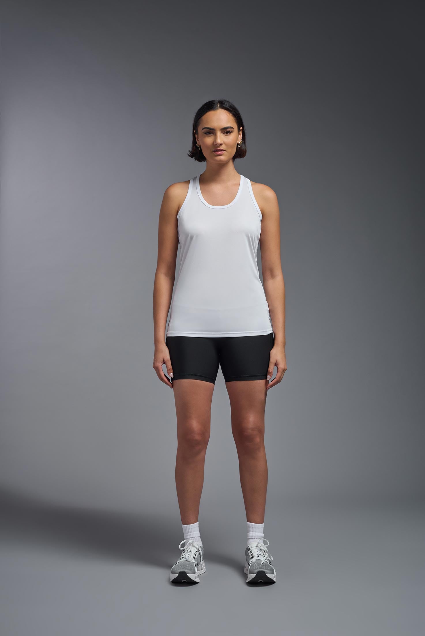 A female model wearing the women's DriMax Endurance Racerback Tank in the color white, made out of 100% polyester, showing the relaxed fit from a front view. This tank is quick-drying, moisture-wicking, with up to 30 UV protection and made in the USA. #white