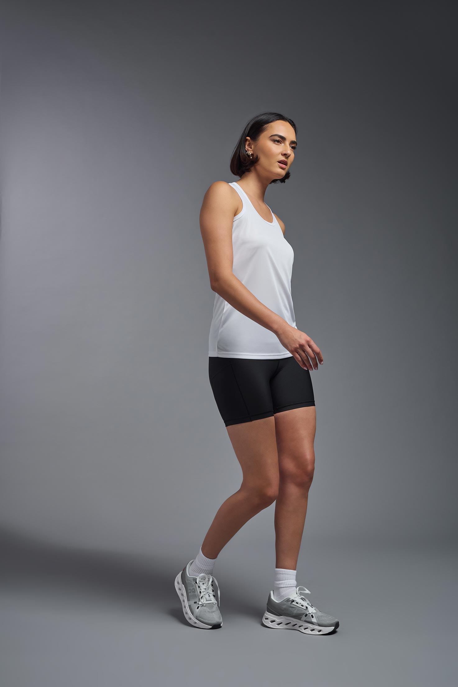 A female model wearing the women's DriMax Endurance Racerback Tank in the color white, made out of 100% polyester, showing the relaxed fit from a side view. This tank is quick-drying, moisture-wicking, with up to 30 UV protection and made in the USA. #white