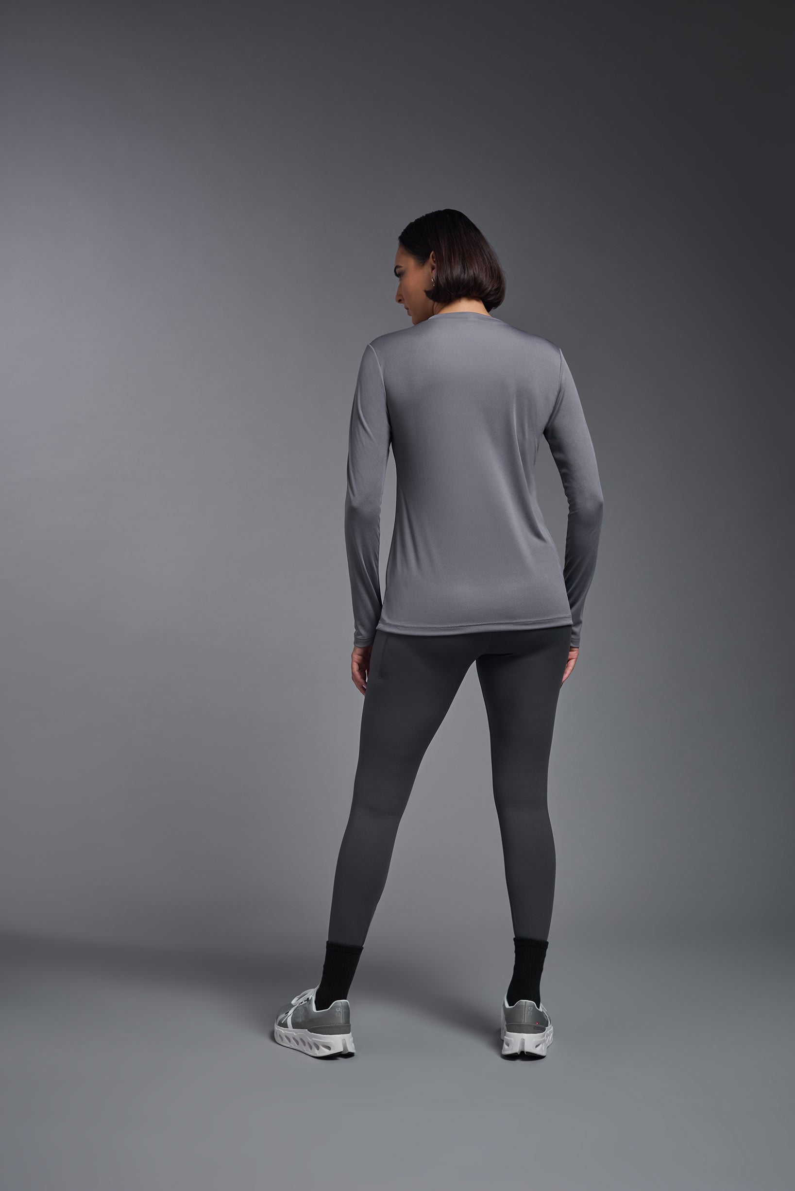 A female model wearing the women's DriMax V-Neck Long Sleeve Expert Tee  in the color steel gray, made out of 100% polyester, showing the relaxed fit from a back view. This shirt is quick-drying, moisture-wicking, with up to 30 UV protection and made in the USA. #steel