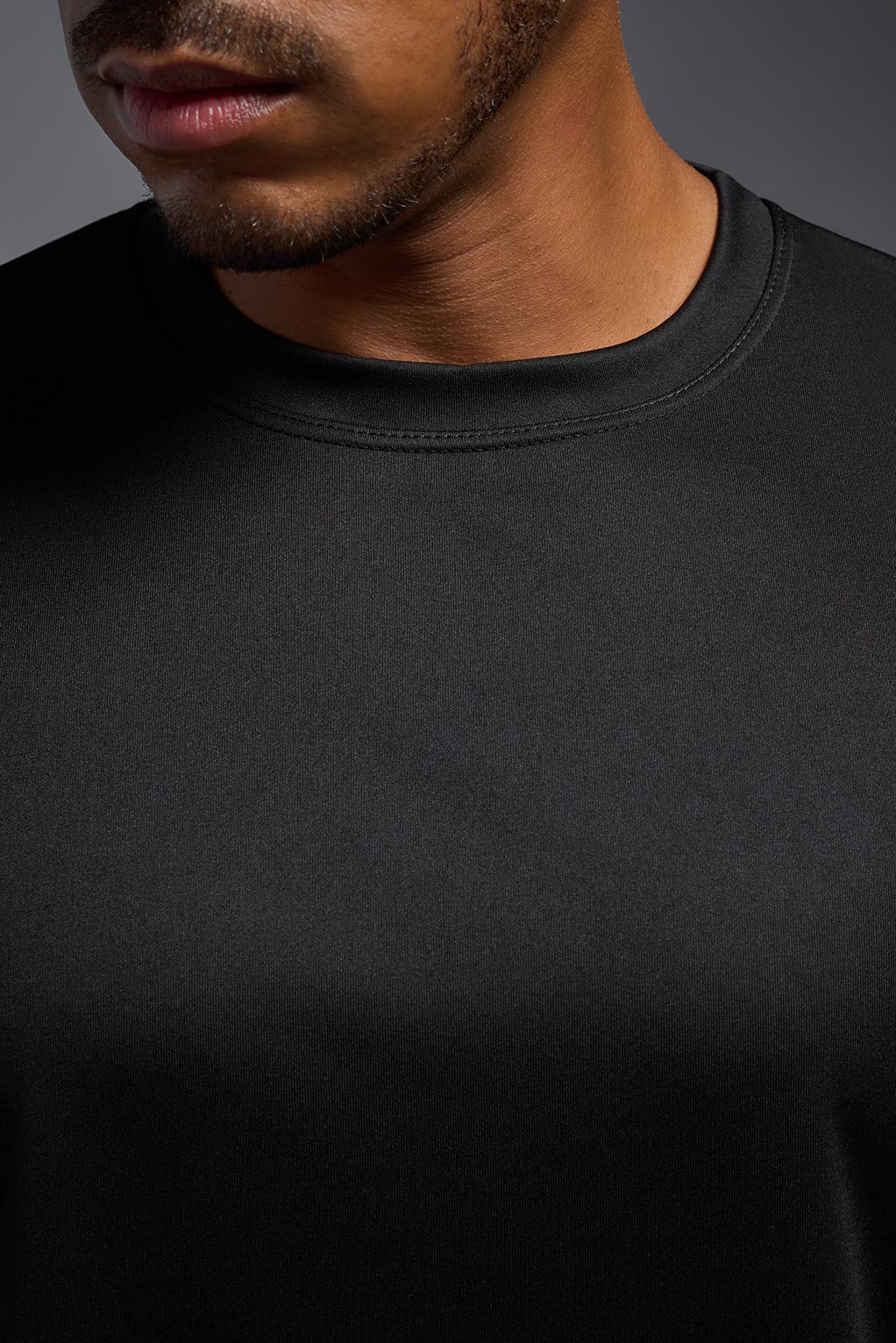 A male model wearing the men's DriMax Expert Tee in the color black, made out of 100% polyester, showing the relaxed fit from a close-up collar view. This shirt is moisture-wicking, quick-drying, and made in the USA. #black
