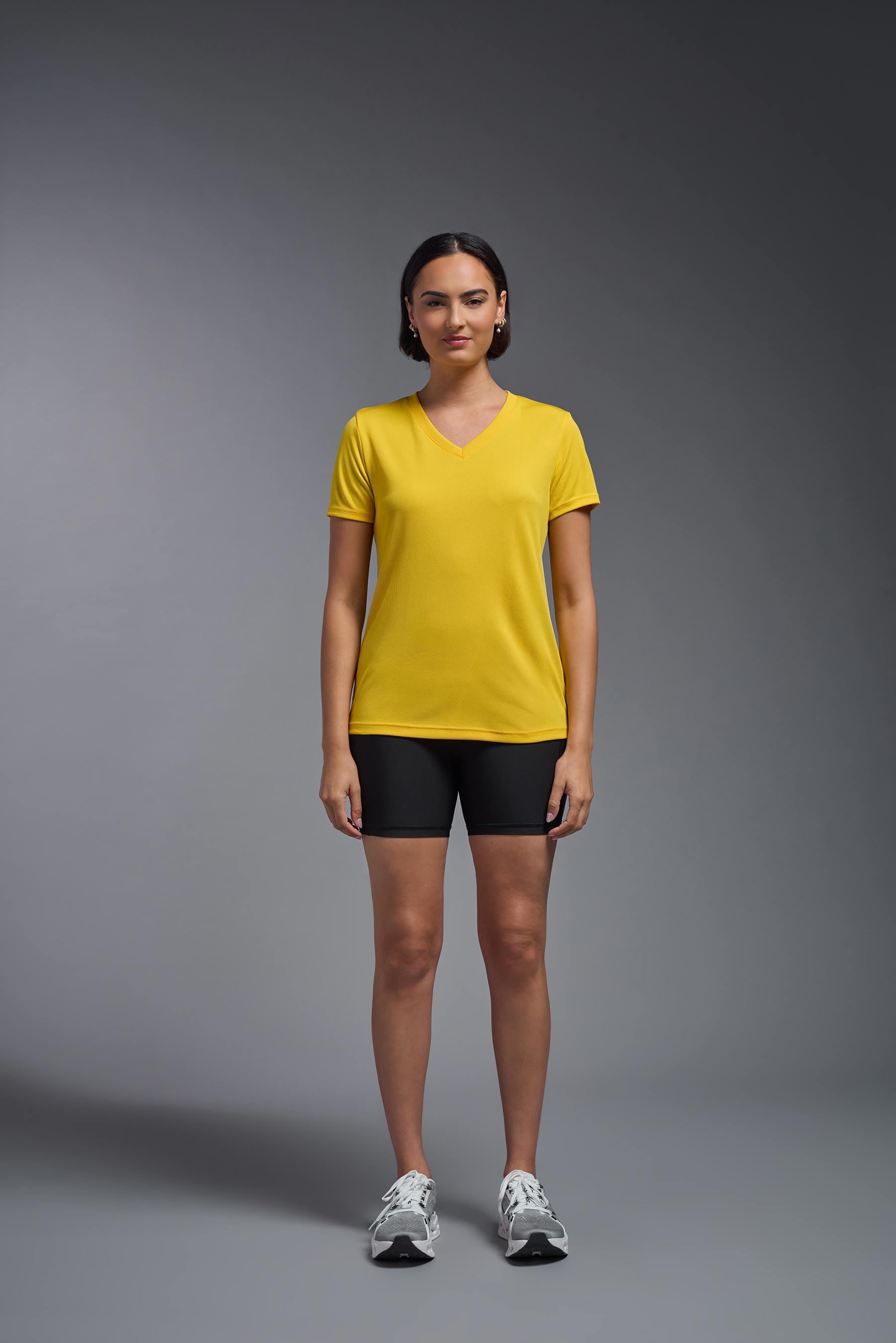 A female model wearing the women's Oxymesh V-neck Tech Tee Imported Version in the color gold, made out of 100% polyester, showing the relaxed fit from a front view. This shirt is breathable, quick-drying, moisture-wicking, and Imported for a better price on bulk apparel. #gold
