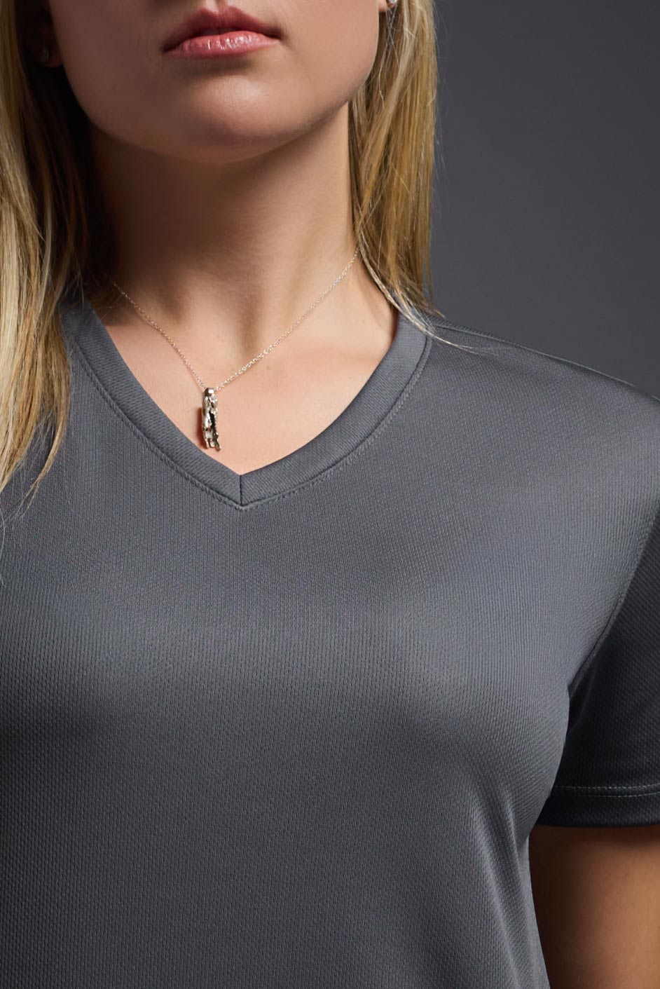 A female model wearing the women's Oxymesh V-neck Tech Tee Imported Version in the color graphite, made out of 100% polyester, showing the relaxed fit from a close-up collar view. This shirt is breathable, quick-drying, moisture-wicking, and Imported for a better price on bulk apparel. #graphite-gray