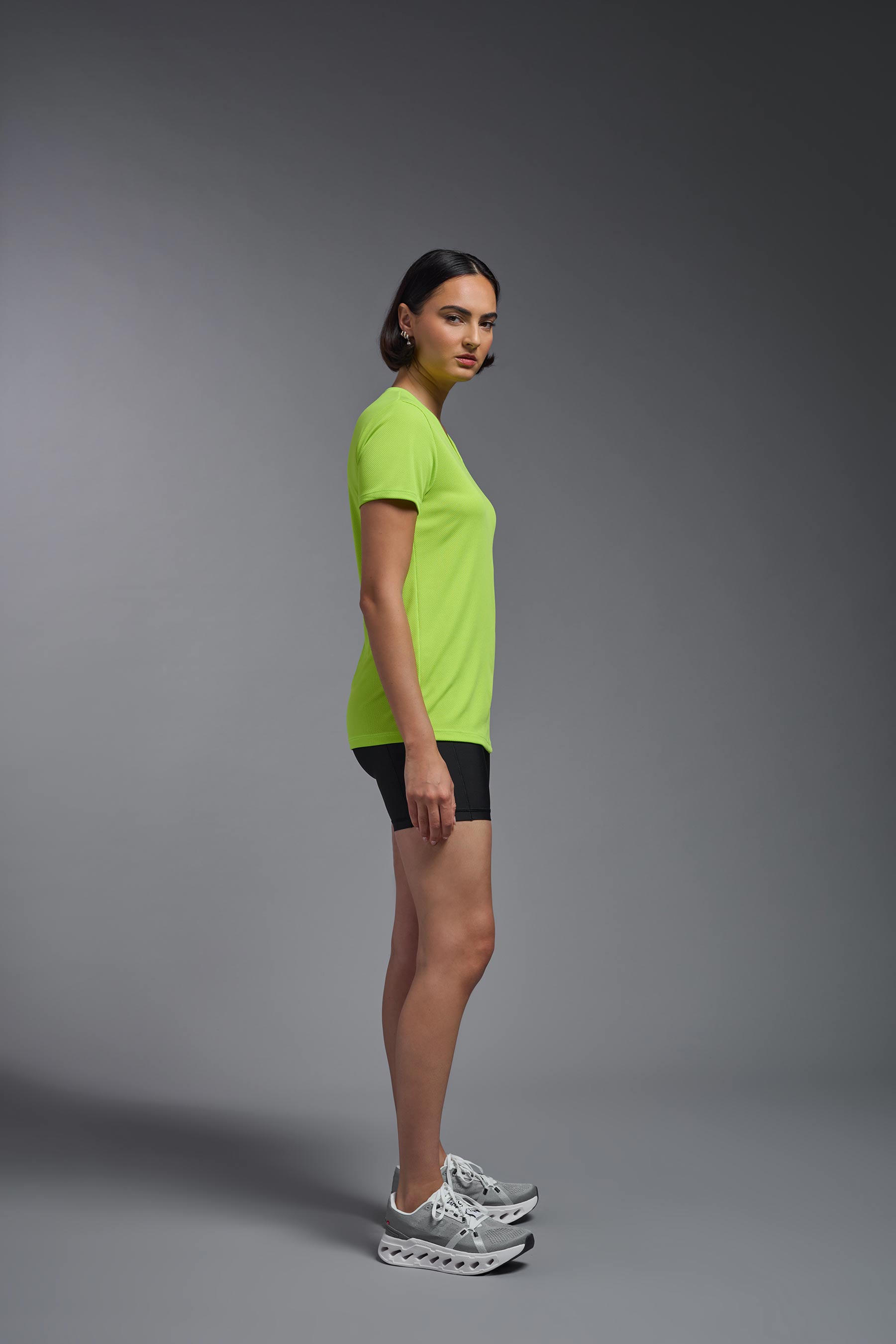 A female model wearing the women's Oxymesh V-neck Tech Tee Imported Version in the color key lime, made out of 100% polyester, showing the relaxed fit from a side view. This shirt is breathable, quick-drying, moisture-wicking, and Imported for a better price on bulk apparel. #key-lime