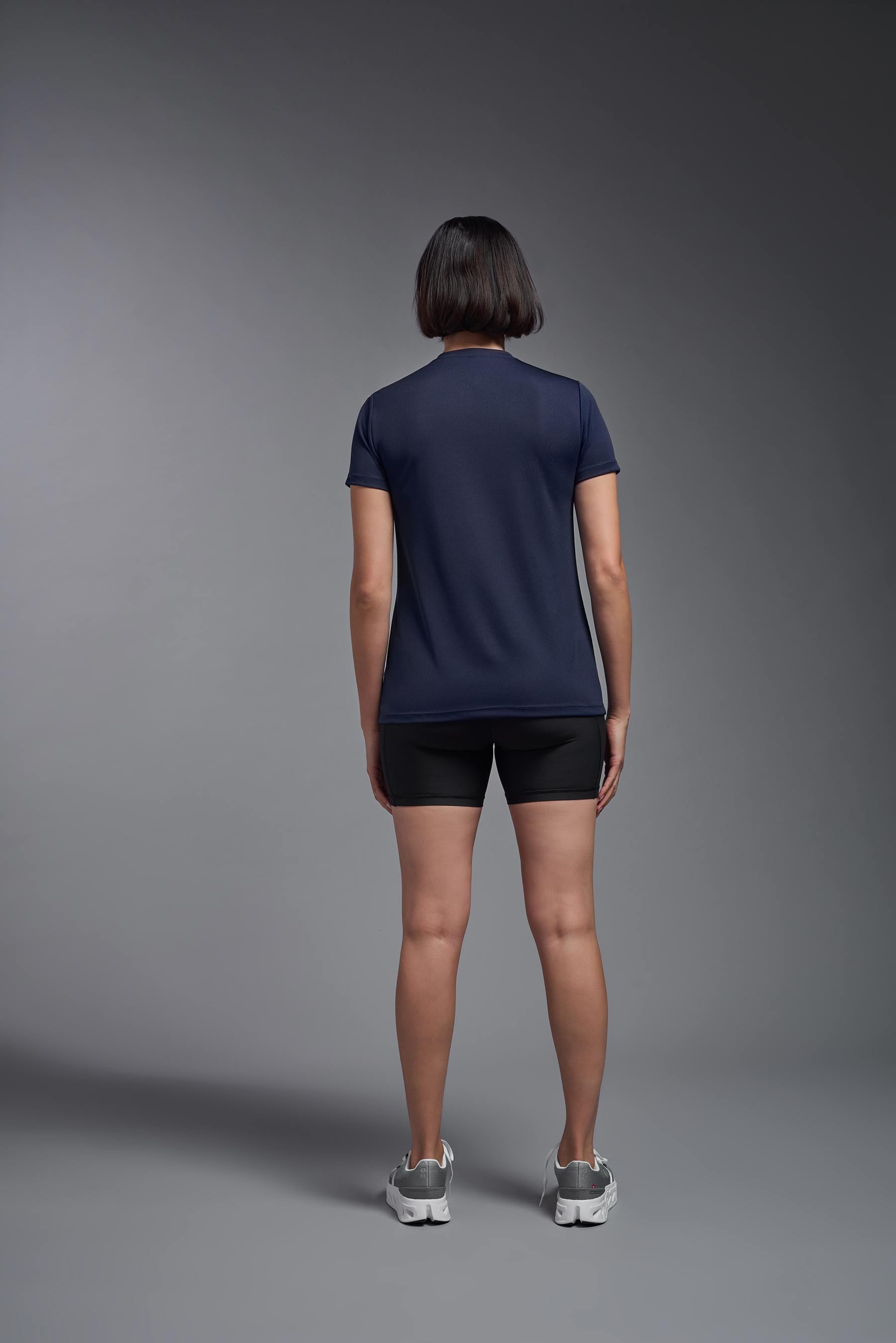 A female model wearing the women's Oxymesh V-neck Tech Tee Imported Version in the color navy, made out of 100% polyester, showing the relaxed fit from a back view. This shirt is breathable, quick-drying, moisture-wicking, and Imported for a better price on bulk apparel. #navy-blue