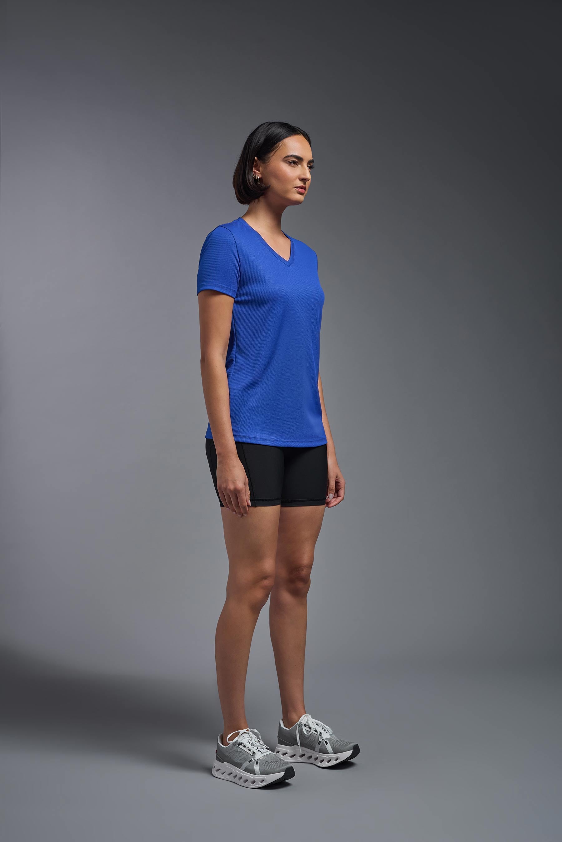 A female model wearing the women's Oxymesh V-neck Tech Tee Imported Version in the color royal blue, made out of 100% polyester, showing the relaxed fit from a side view. This shirt is breathable, quick-drying, moisture-wicking, and Imported for a better price on bulk apparel. #royal-blue