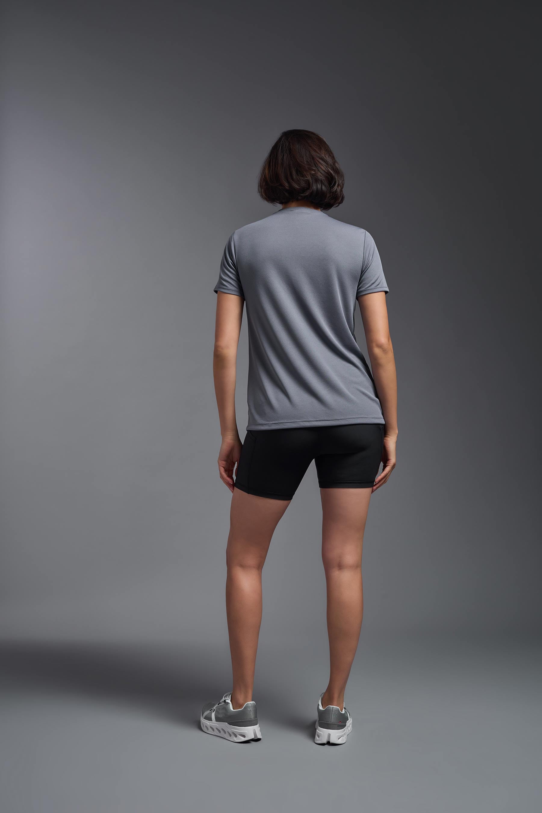 A female model wearing the women's Oxymesh V-neck Tech Tee Imported Version in the color steel gray, made out of 100% polyester, showing the relaxed fit from a back view. This shirt is breathable, quick-drying, moisture-wicking, and Imported for a better price on bulk apparel. #steel-gray