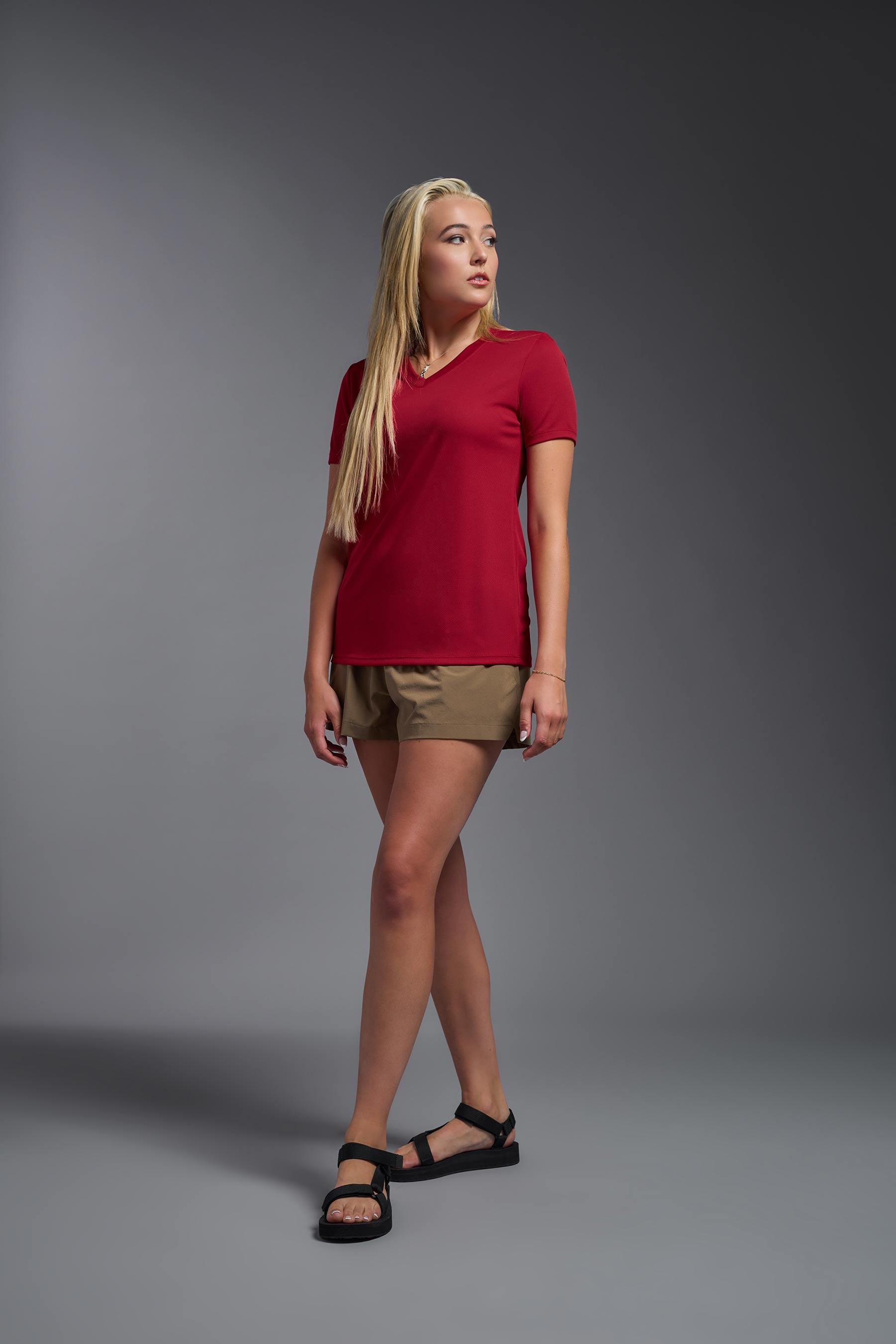 A female model wearing the women's Oxymesh V-neck Tech Tee Imported Version in the color true red, made out of 100% polyester, showing the relaxed fit from a side view. This shirt is breathable, quick-drying, moisture-wicking, and Imported for a better price on bulk apparel. #true-red