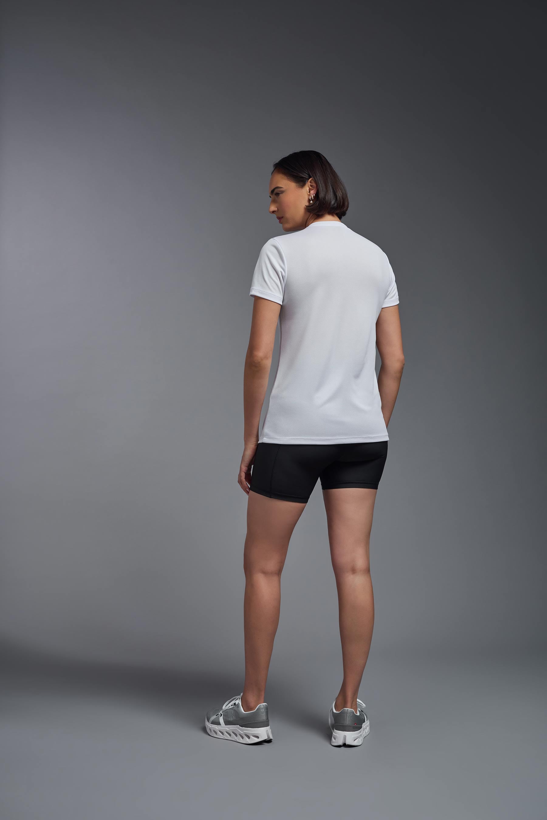 A female model wearing the women's Oxymesh V-neck Tech Tee Imported Version in the color white, made out of 100% polyester, showing the relaxed fit from a back view. This shirt is breathable, quick-drying, moisture-wicking, and Imported for a better price on bulk apparel. #white