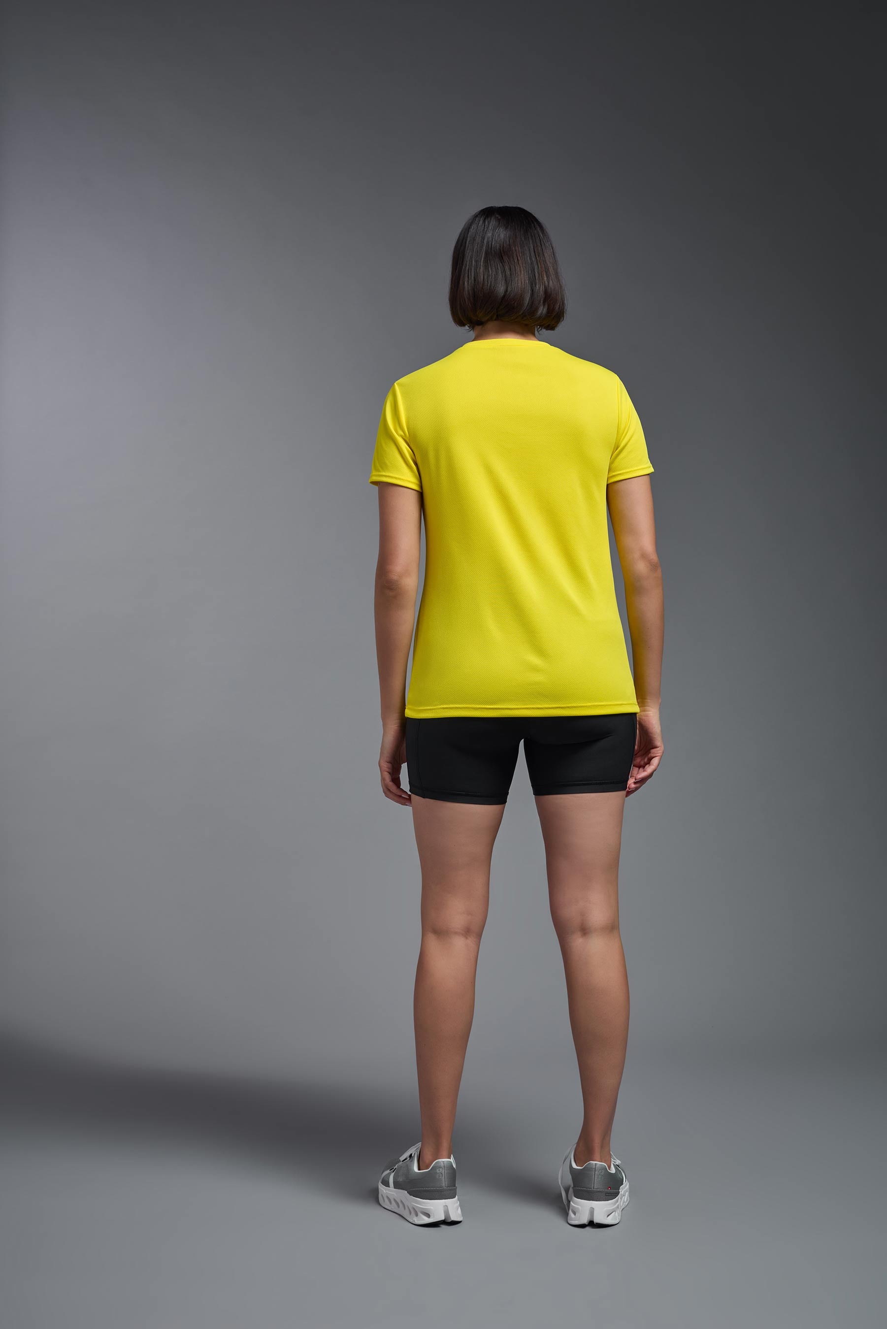 A female model wearing the women's Oxymesh V-neck Tech Tee in the color bright yellow, made out of 100% polyester, showing the relaxed fit from a back view. This shirt is breathable, wicks away moisture, quick-dries, and is made in the USA. #bright-yellow
