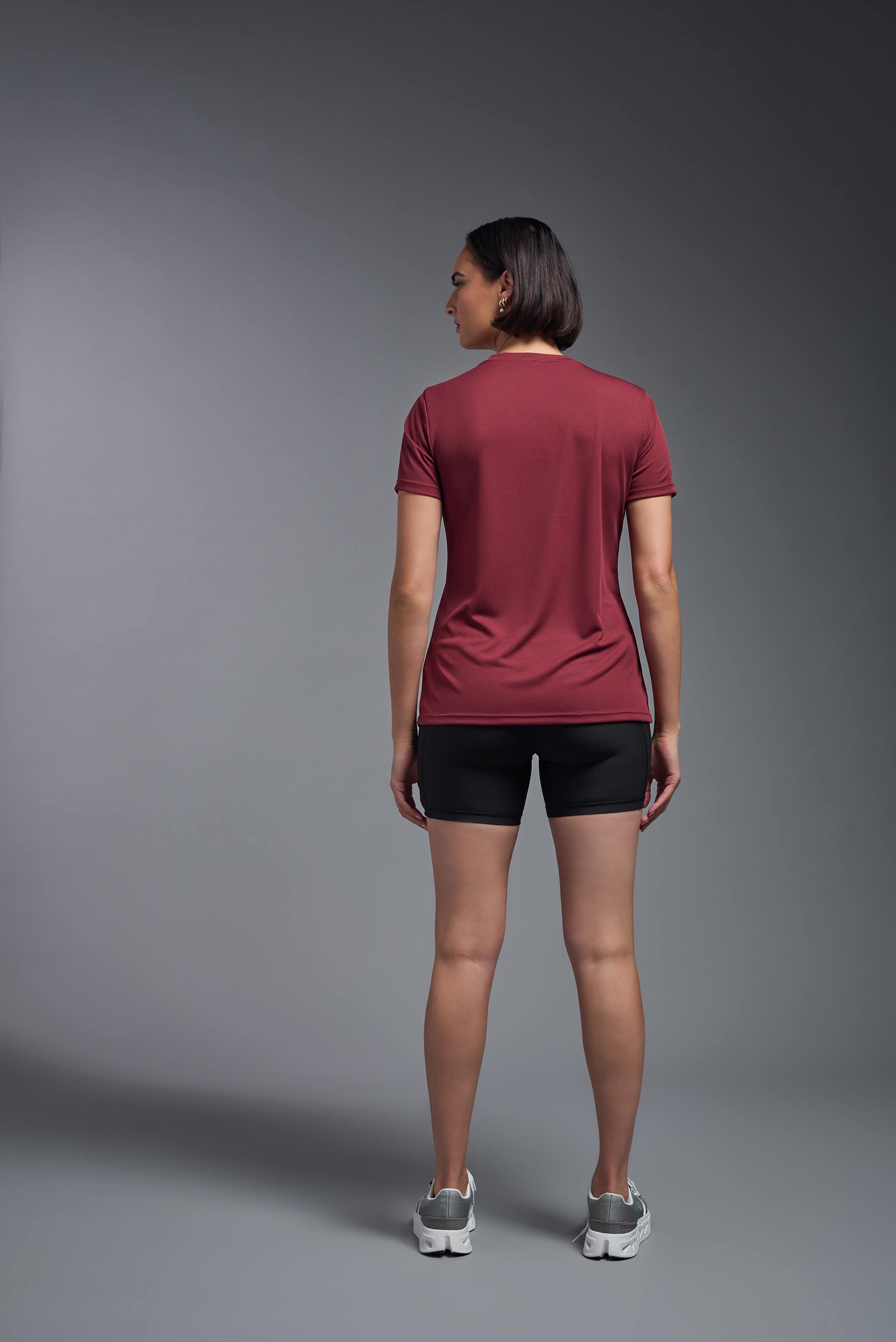 A female model wearing the women's Oxymesh V-neck Tech Tee in the color cardinal, made out of 100% polyester, showing the relaxed fit from a back view. This shirt is breathable, wicks away moisture, quick-dries, and is made in the USA. #cardinal-red