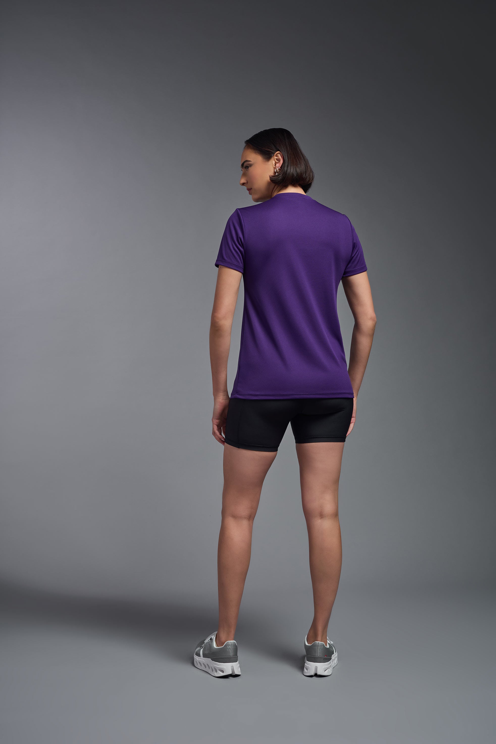 A female model wearing the women's Oxymesh V-neck Tech Tee in the color dark purple, made out of 100% polyester, showing the relaxed fit from a back view. This shirt is breathable, wicks away moisture, quick-dries, and is made in the USA. #dark-purple