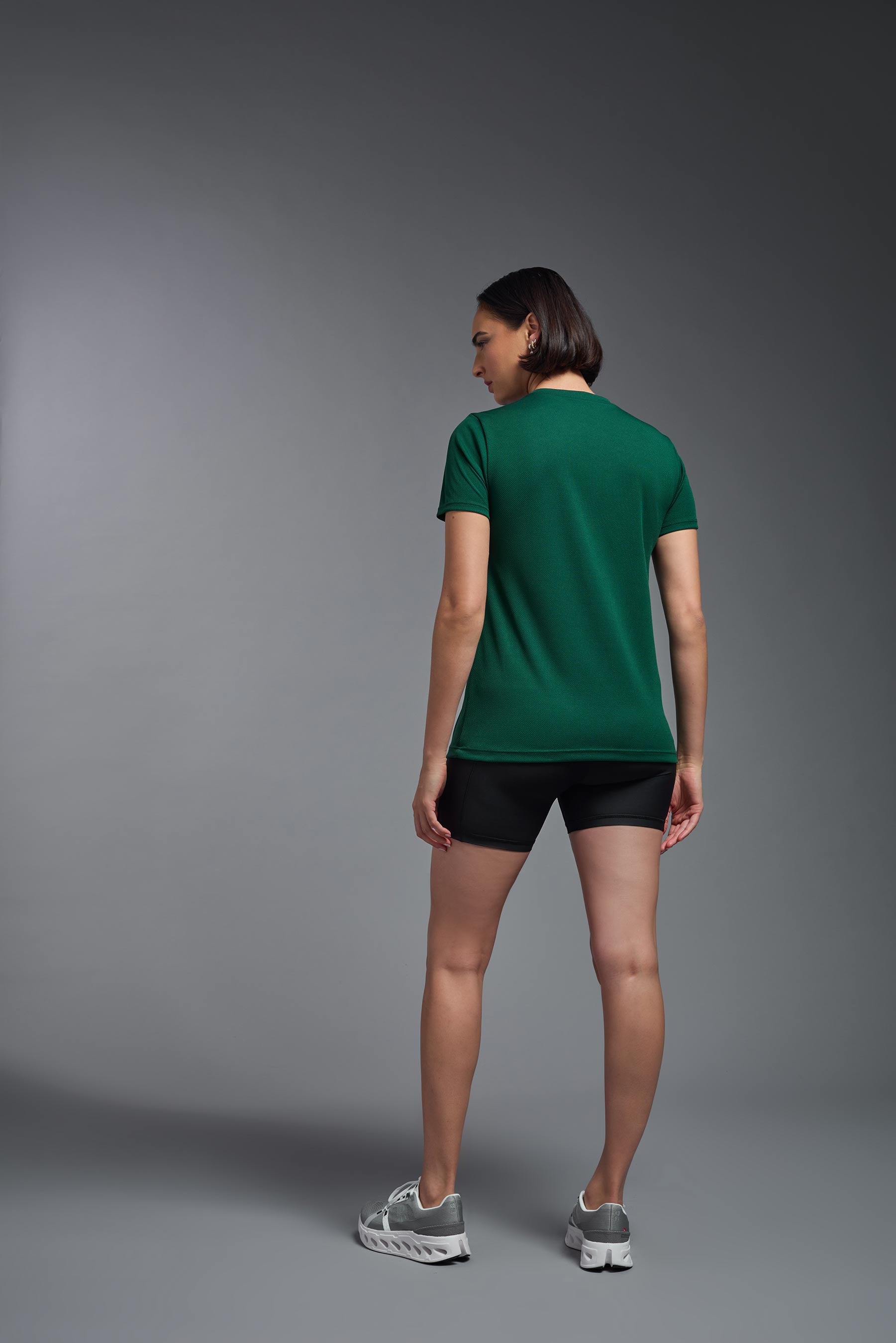 A female model wearing the women's Oxymesh V-neck Tech Tee in the color forest green, made out of 100% polyester, showing the relaxed fit from a back view. This shirt is breathable, wicks away moisture, quick-dries, and is made in the USA. #forest-green