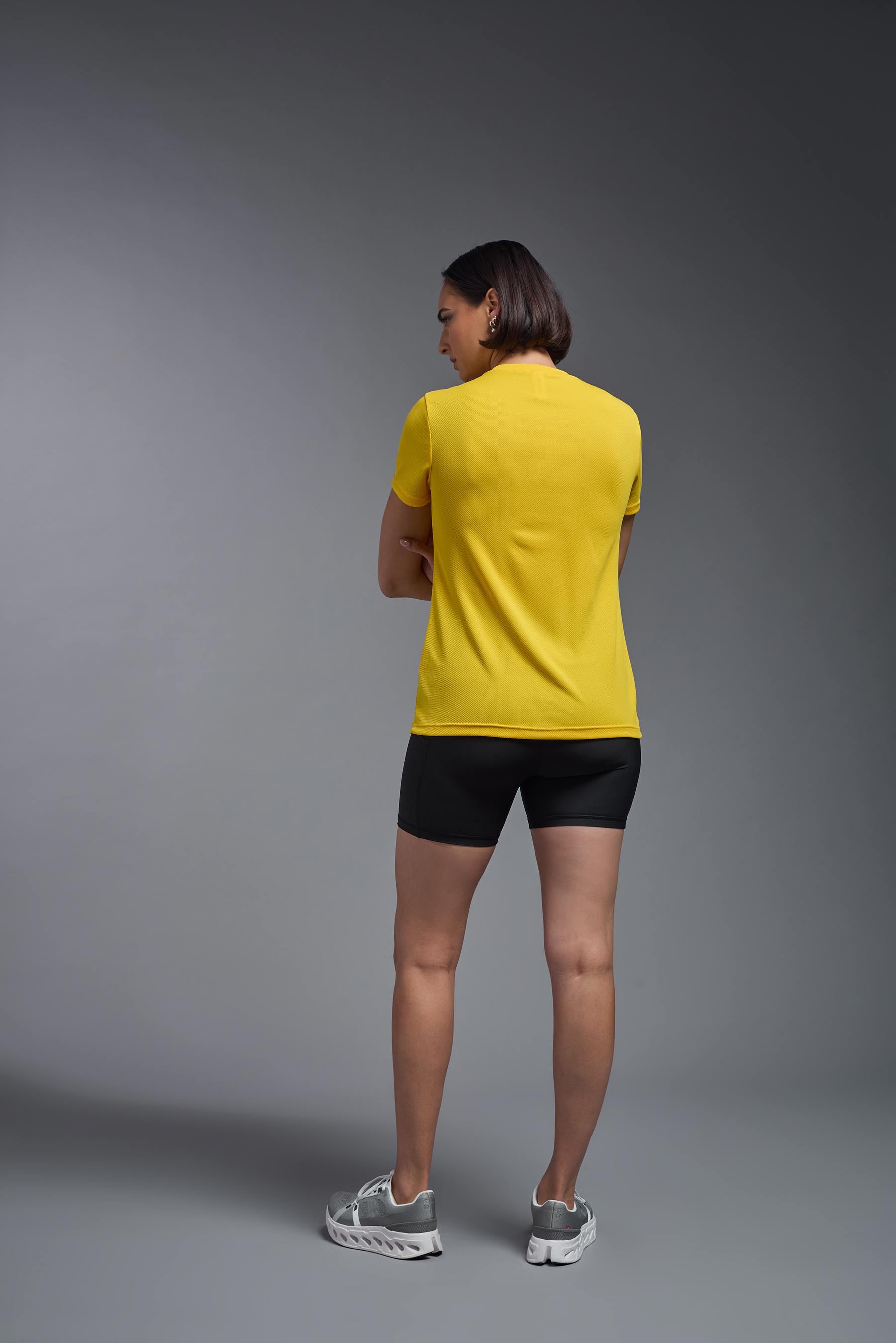 A female model wearing the women's Oxymesh V-neck Tech Tee in the color gold, made out of 100% polyester, showing the relaxed fit from a back view. This shirt is breathable, wicks away moisture, quick-dries, and is made in the USA. #gold