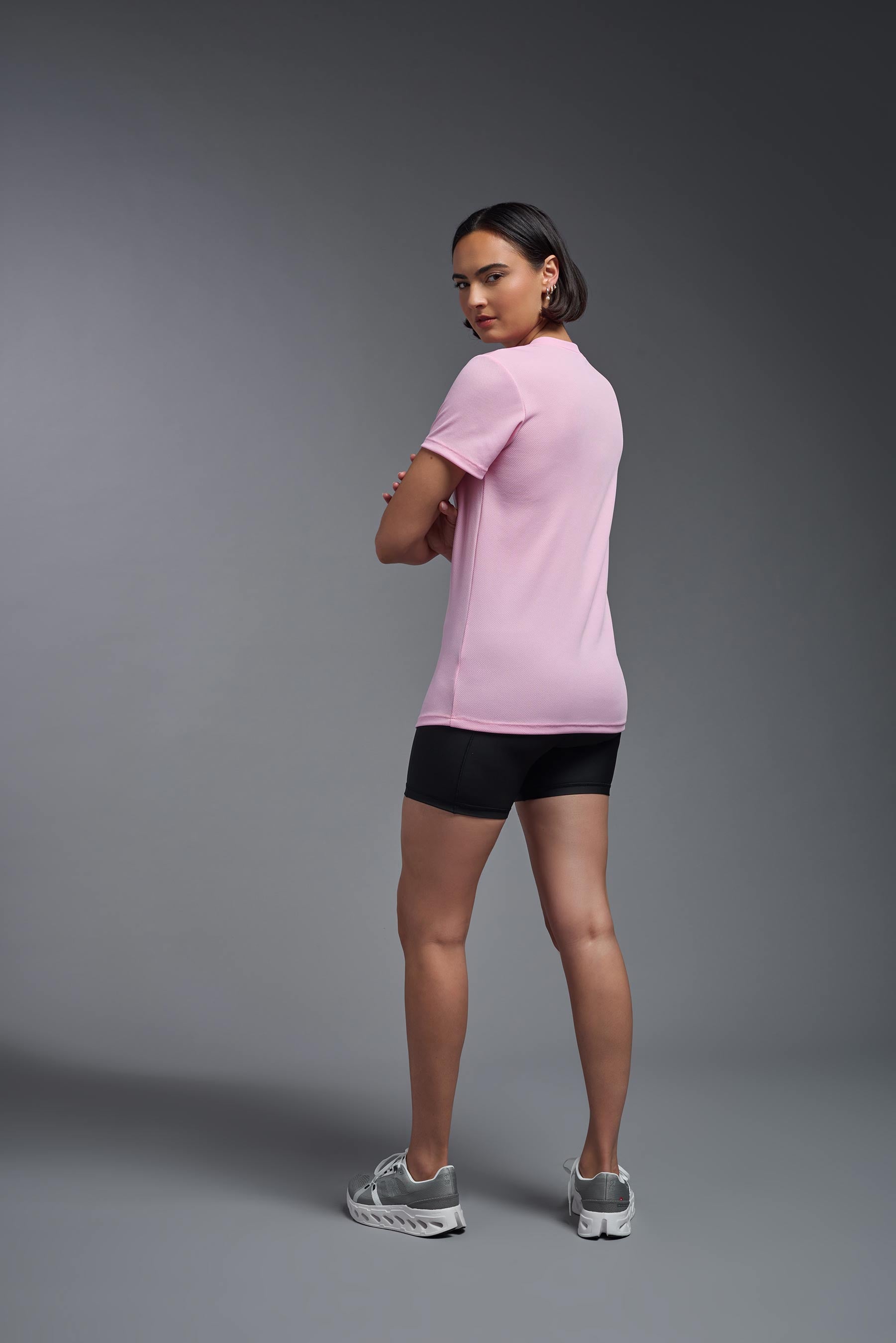 A female model wearing the women's Oxymesh V-neck Tech Tee in the color pink, made out of 100% polyester, showing the relaxed fit from a back view. This shirt is breathable, wicks away moisture, quick-dries, and is made in the USA. #pink