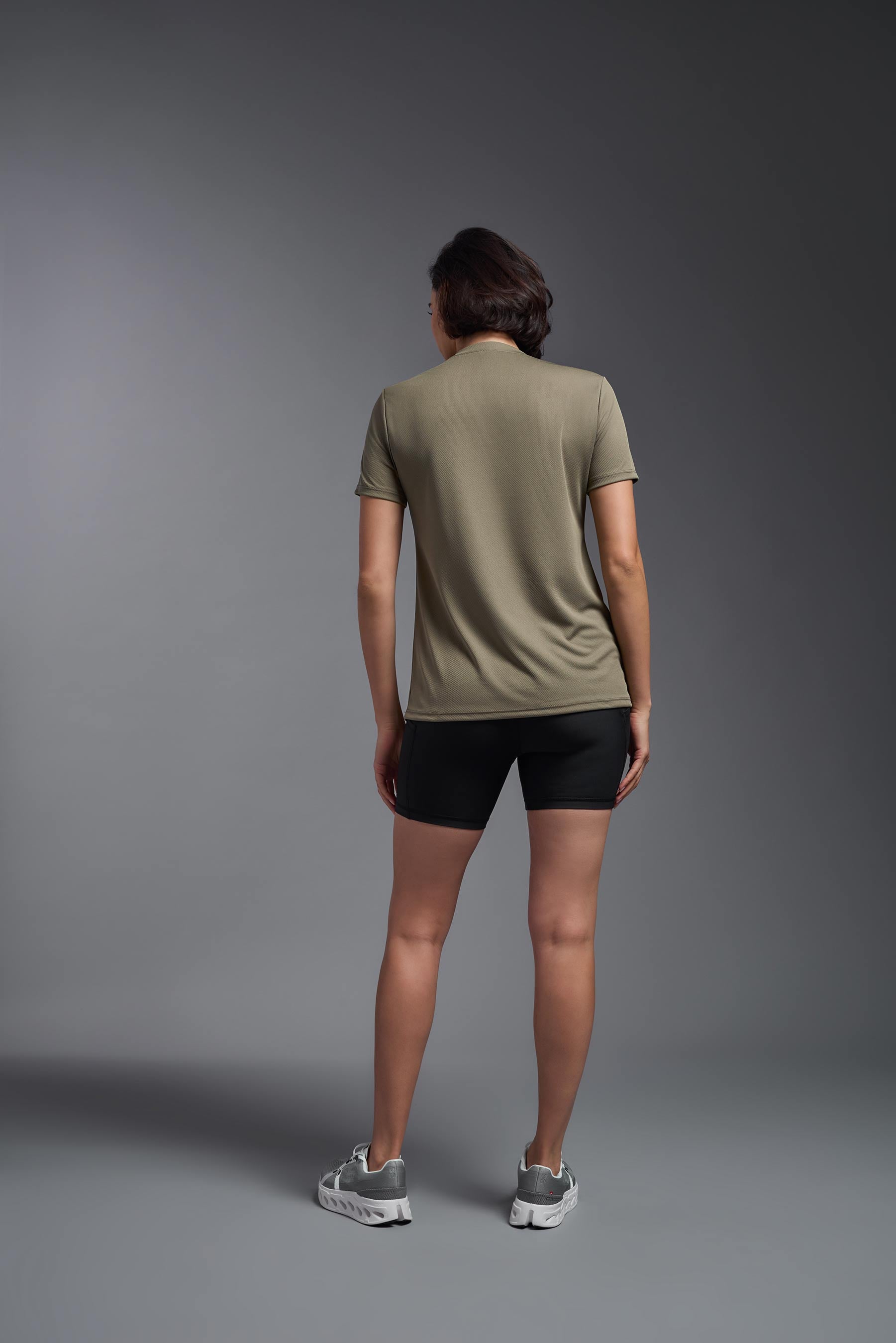 A female model wearing the women's Oxymesh V-neck Tech Tee in the color tan, made out of 100% polyester, showing the relaxed fit from a back view. This shirt is breathable, wicks away moisture, quick-dries, and is made in the USA. #tan