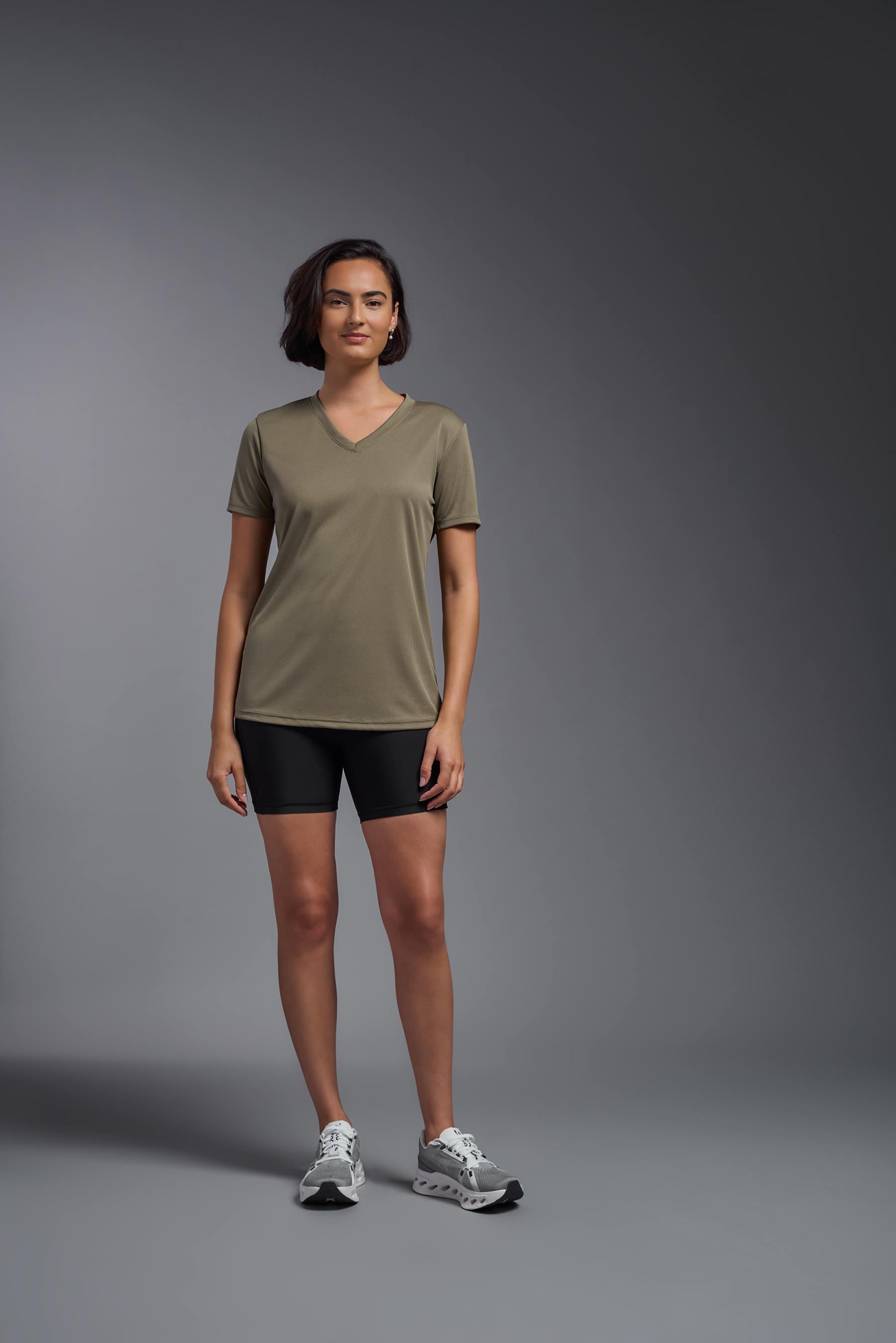 A female model wearing the women's Oxymesh V-neck Tech Tee in the color tan, made out of 100% polyester, showing the relaxed fit from a front view. This shirt is breathable, wicks away moisture, quick-dries, and is made in the USA. #tan