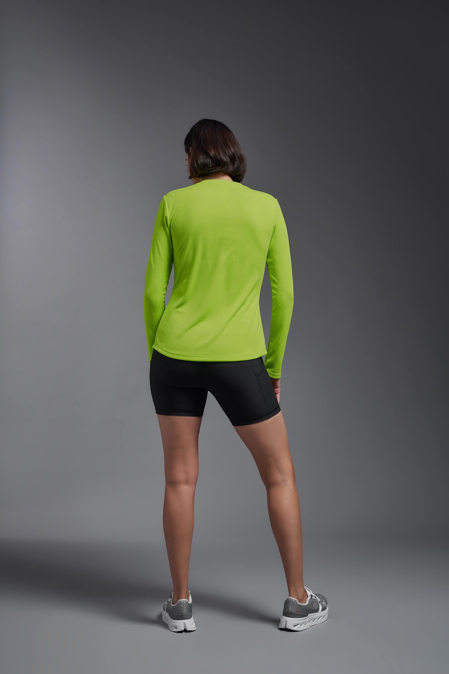A female model wearing the women's Oxymesh Long Sleeve Tech Tee Imported Version in the color key lime, made out of 100% polyester, showing the relaxed fit from a back view. This shirt is breathable, quick-drying, moisture-wicking, and Imported for a better price on bulk apparel. #key-lime