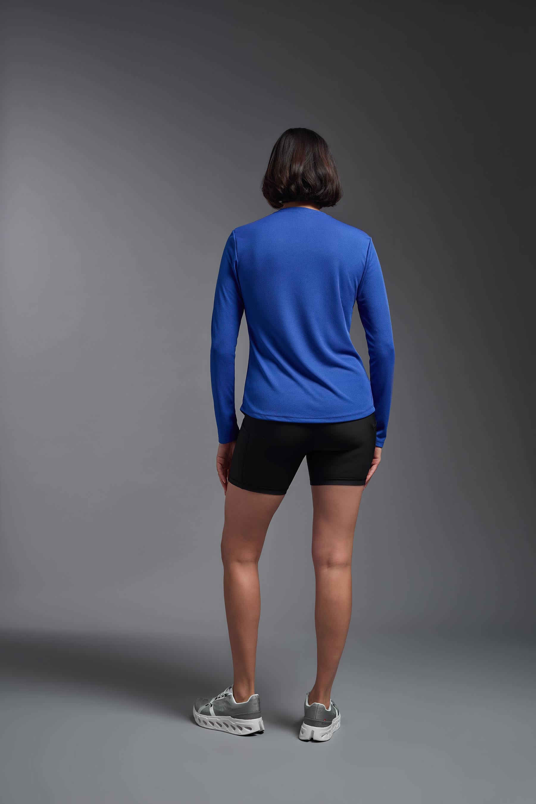 A female model wearing the women's Oxymesh Long Sleeve Tech Tee Imported Version in the color royal blue, made out of 100% polyester, showing the relaxed fit from a back view. This shirt is breathable, quick-drying, moisture-wicking, and Imported for a better price on bulk apparel. #royal-blue