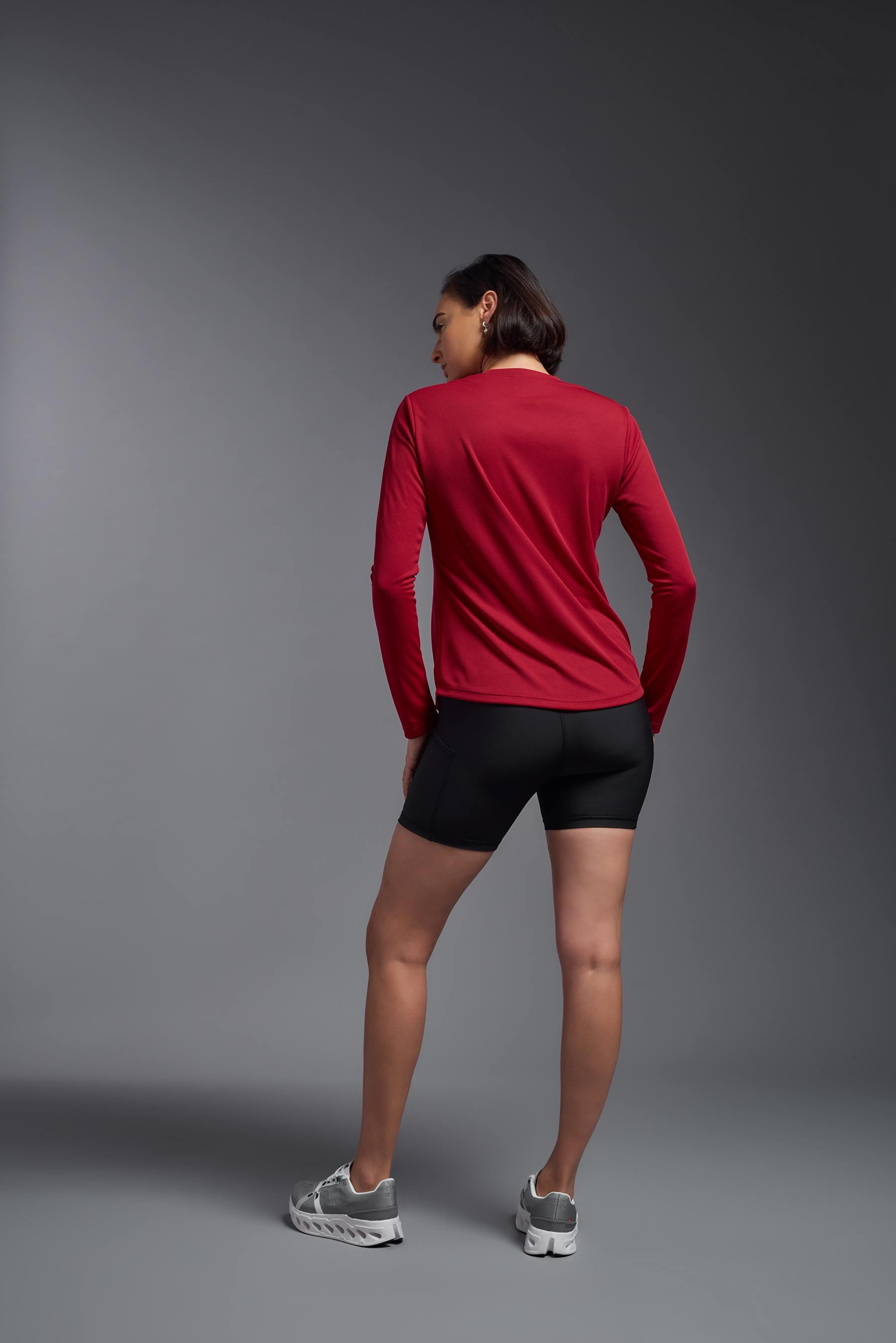 A female model wearing the women's Oxymesh Long Sleeve Tech Tee Imported Version in the color true red, made out of 100% polyester, showing the relaxed fit from a back view. This shirt is breathable, quick-drying, moisture-wicking, and Imported for a better price on bulk apparel. #true-red