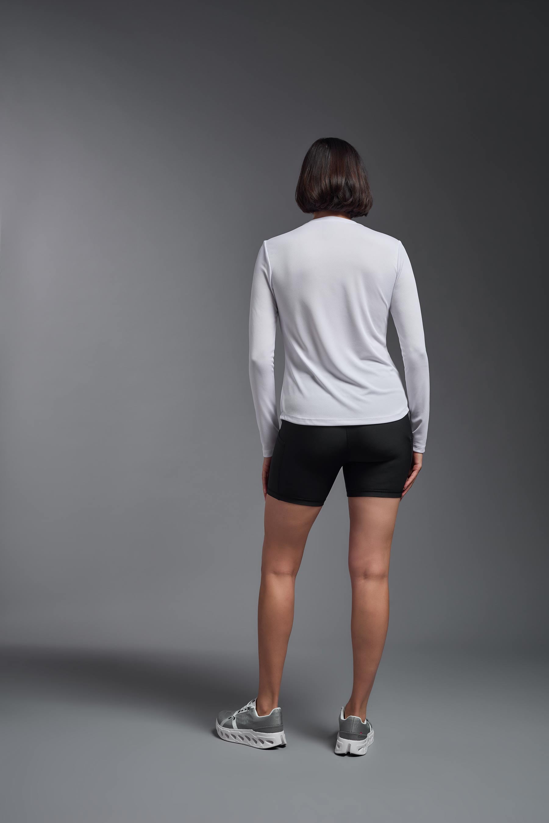 A female model wearing the women's Oxymesh Long Sleeve Tech Tee Imported Version in the color white, made out of 100% polyester, showing the relaxed fit from a back view. This shirt is breathable, quick-drying, moisture-wicking, and Imported for a better price on bulk apparel. #white