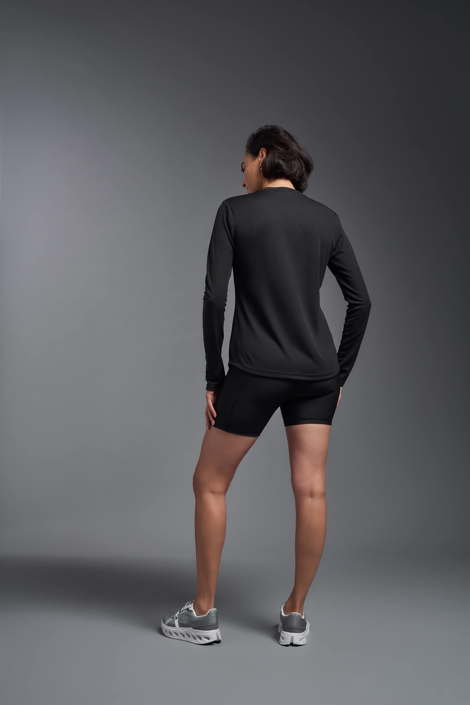 A female model wearing the women's Oxymesh Long Sleeve Tech Tee in the color black, made out of 100% polyester, showing the relaxed fit from a back view. This shirt is breathable, quick-drying, moisture-wicking, and made in the USA. #black