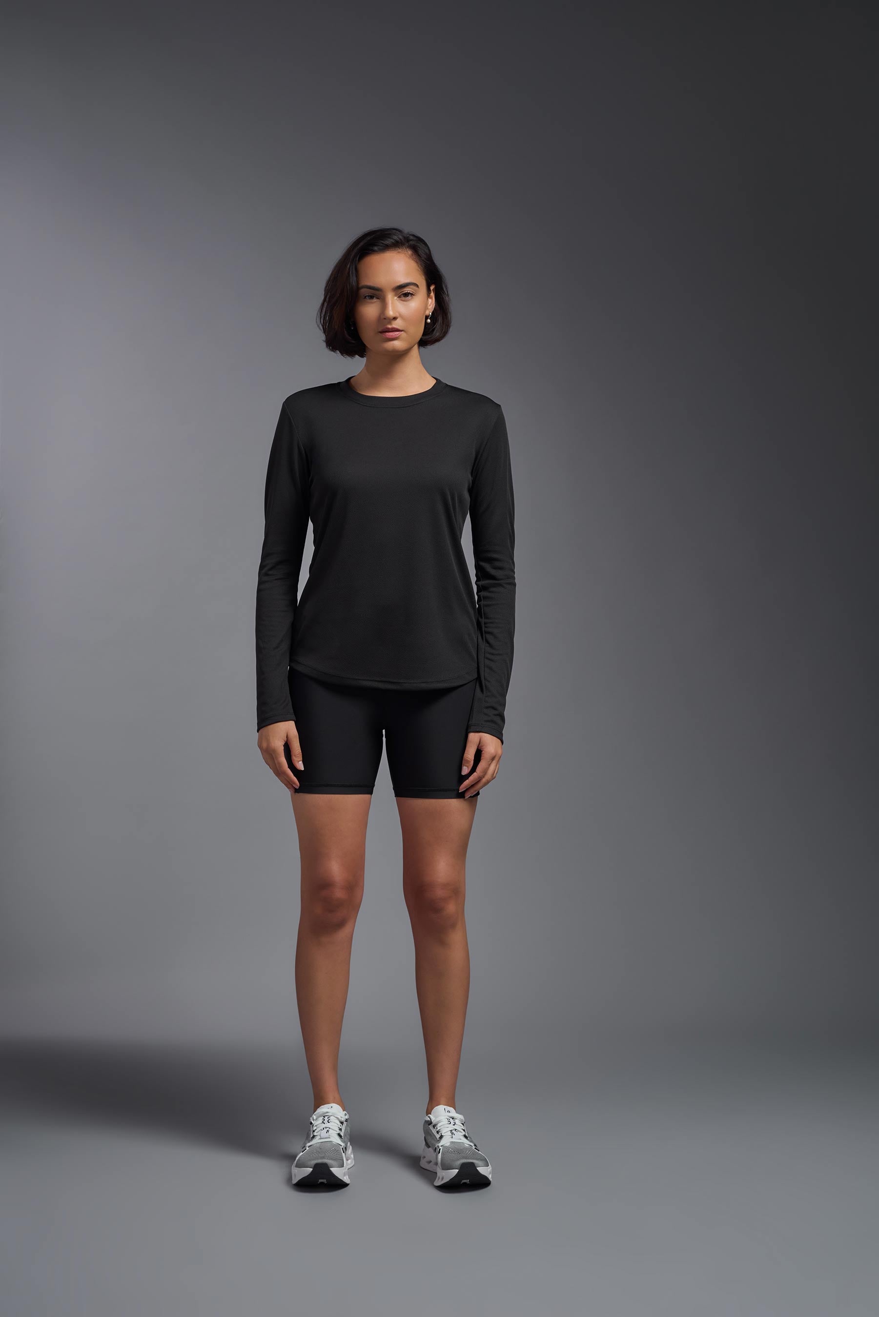 A female model wearing the women's Oxymesh Long Sleeve Tech Tee in the color black, made out of 100% polyester, showing the relaxed fit from a front view. This shirt is breathable, quick-drying, moisture-wicking, and made in the USA. #black
