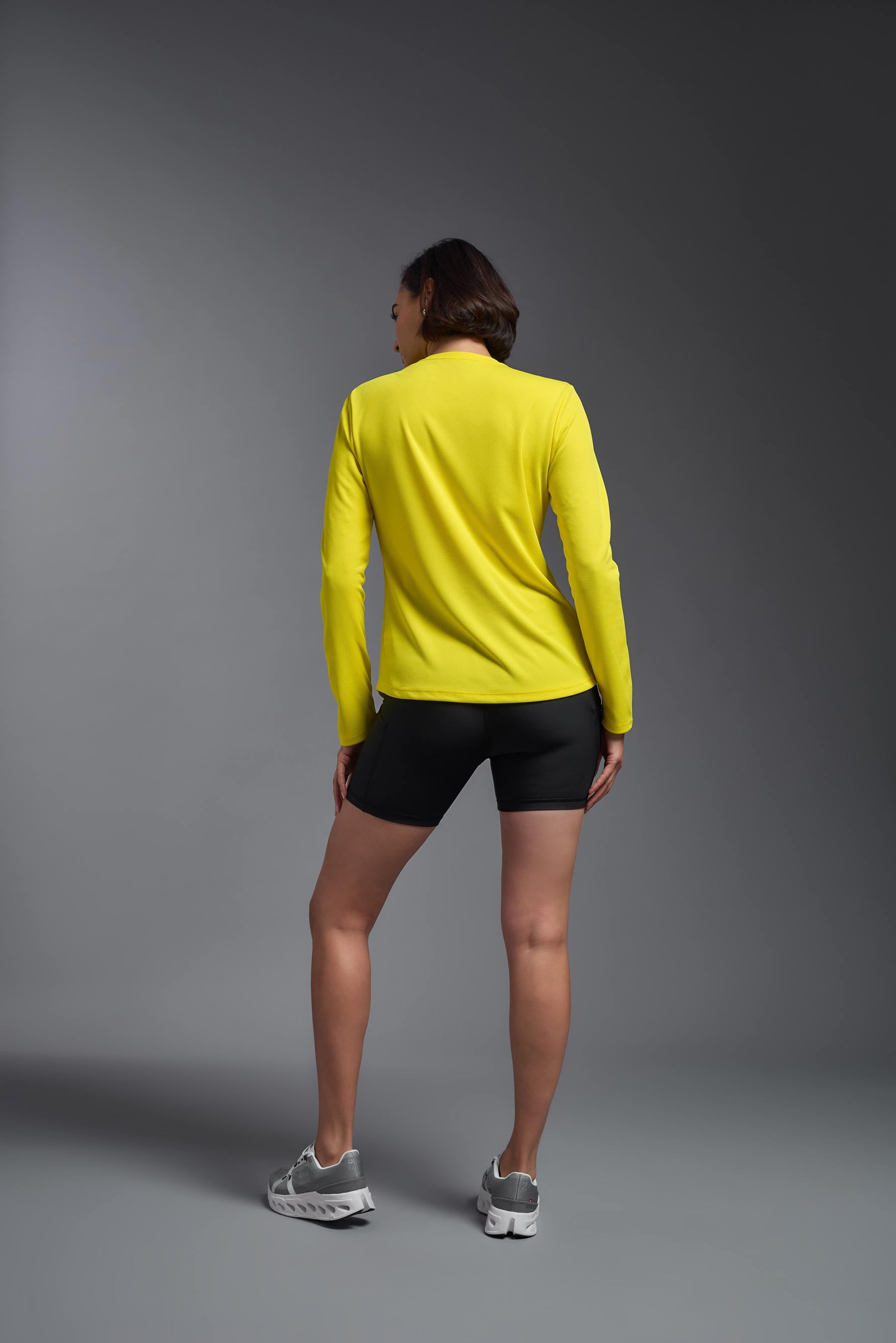 A female model wearing the women's Oxymesh Long Sleeve Tech Tee in the color bright yellow, made out of 100% polyester, showing the relaxed fit from a back view. This shirt is breathable, quick-drying, moisture-wicking, and made in the USA. #bright-yellow