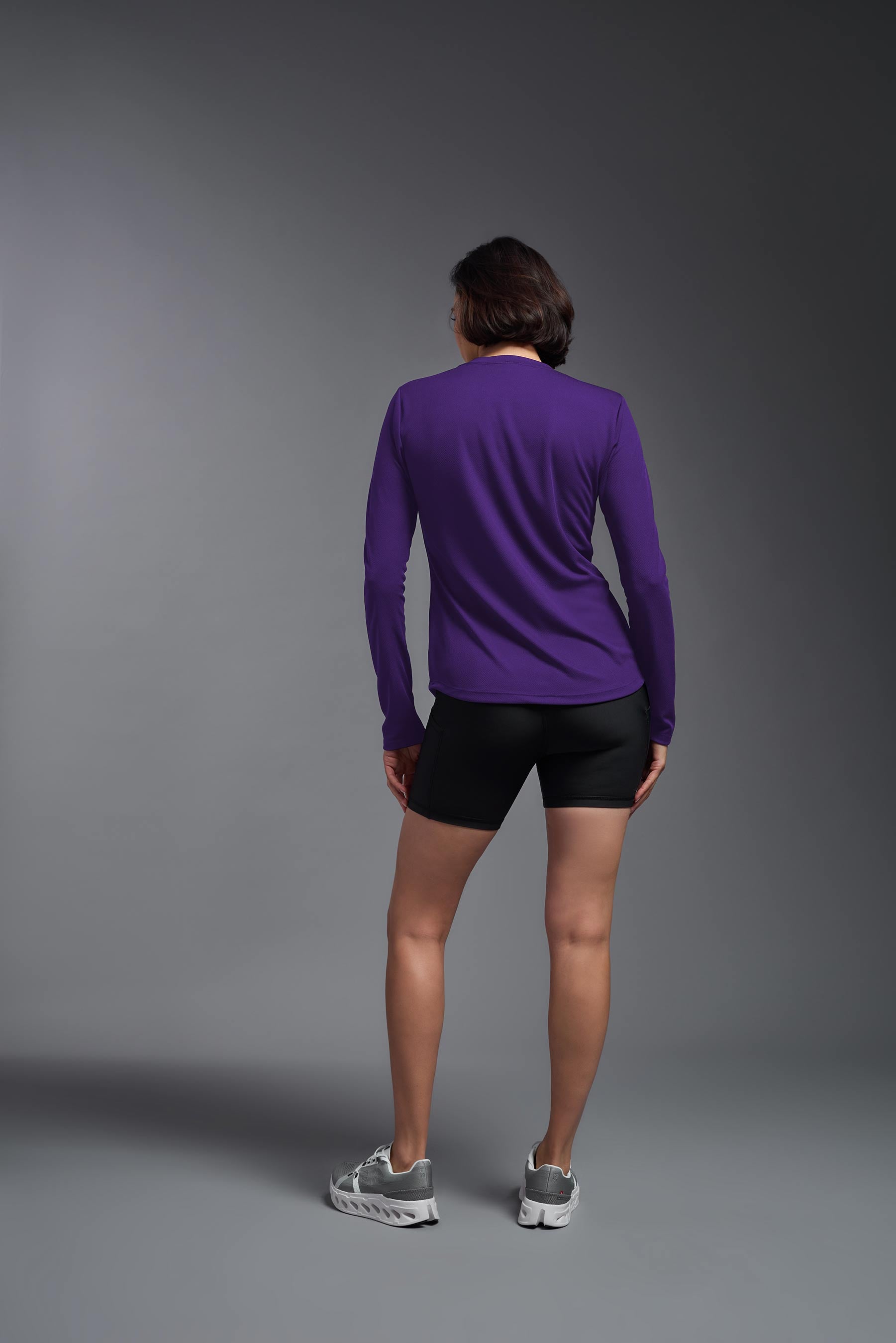 A female model wearing the women's Oxymesh Long Sleeve Tech Tee in the color dark purple, made out of 100% polyester, showing the relaxed fit from a back view. This shirt is breathable, quick-drying, moisture-wicking, and made in the USA. #dark-purple