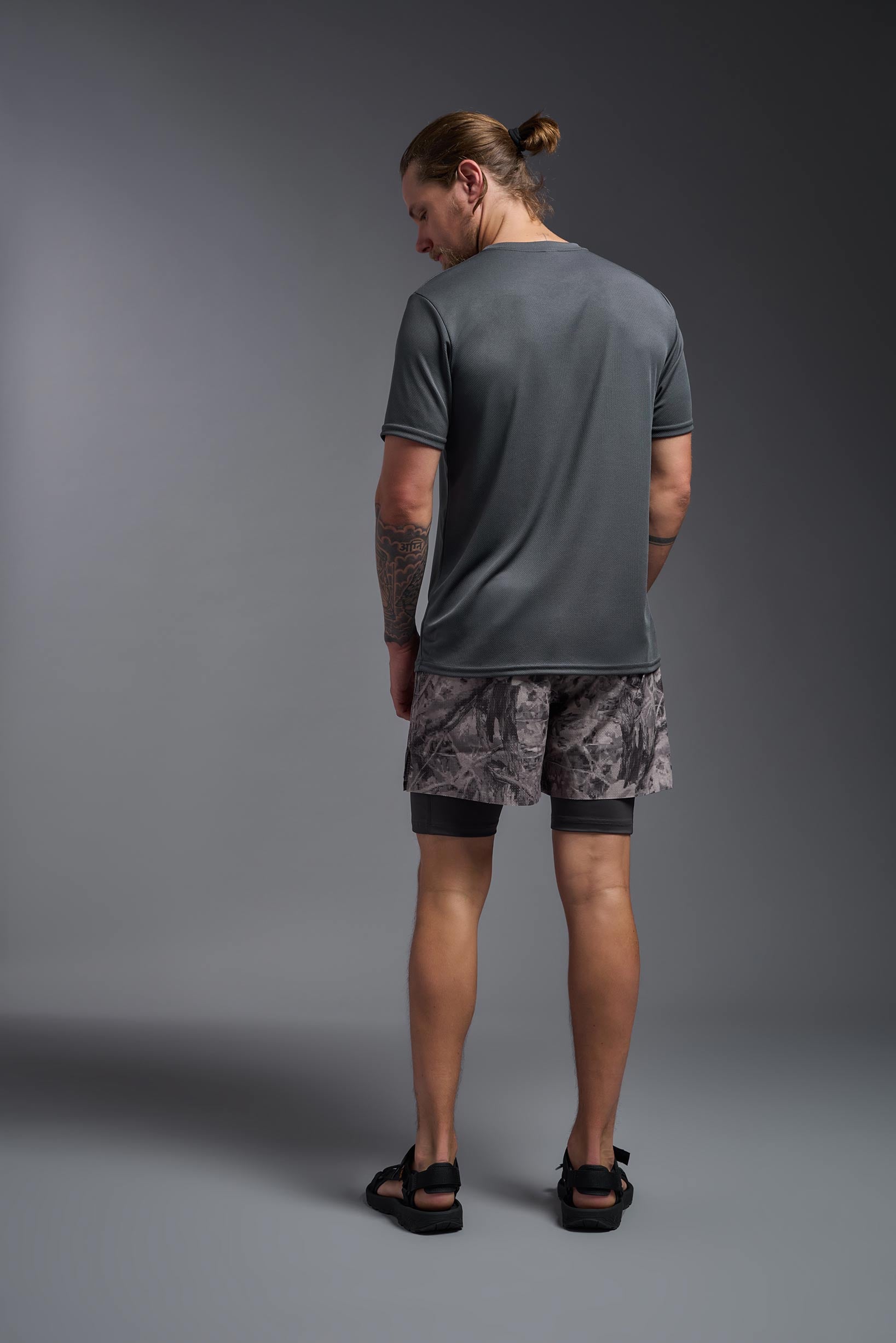 A male model wearing the men's Oxymesh Long Sleeve Tech Tee Imported Version in the color graphite gray, made out of 100% polyester, showing the relaxed fit from a back view. This shirt is breathable, quick-drying, moisture-wicking, and Imported for a better price on bulk apparel. #graphite-gray