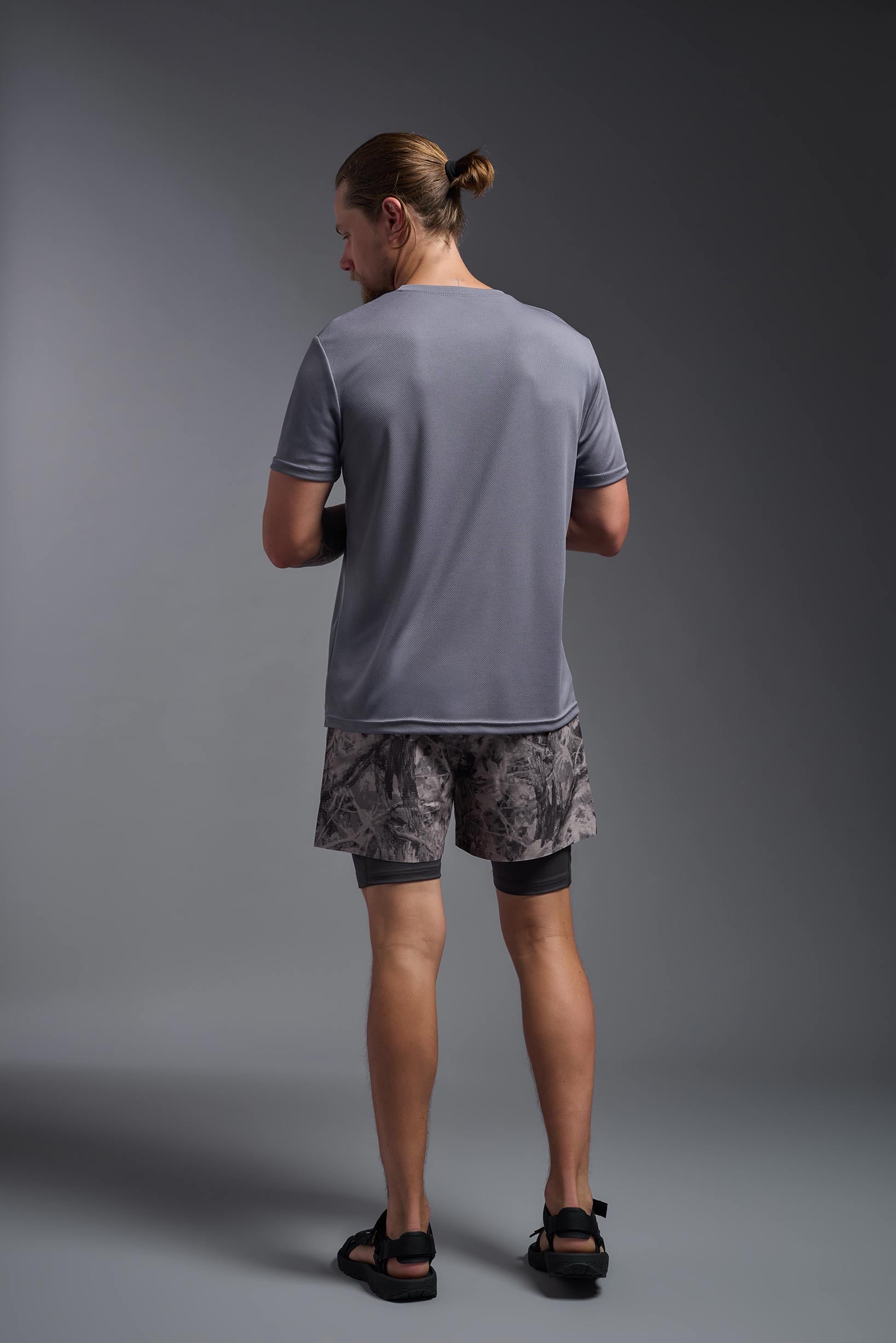 A male model wearing the men's Oxymesh Long Sleeve Tech Tee Imported Version in the color steel gray, made out of 100% polyester, showing the relaxed fit from a back view. This shirt is breathable, quick-drying, moisture-wicking, and Imported for a better price on bulk apparel. #steel-gray