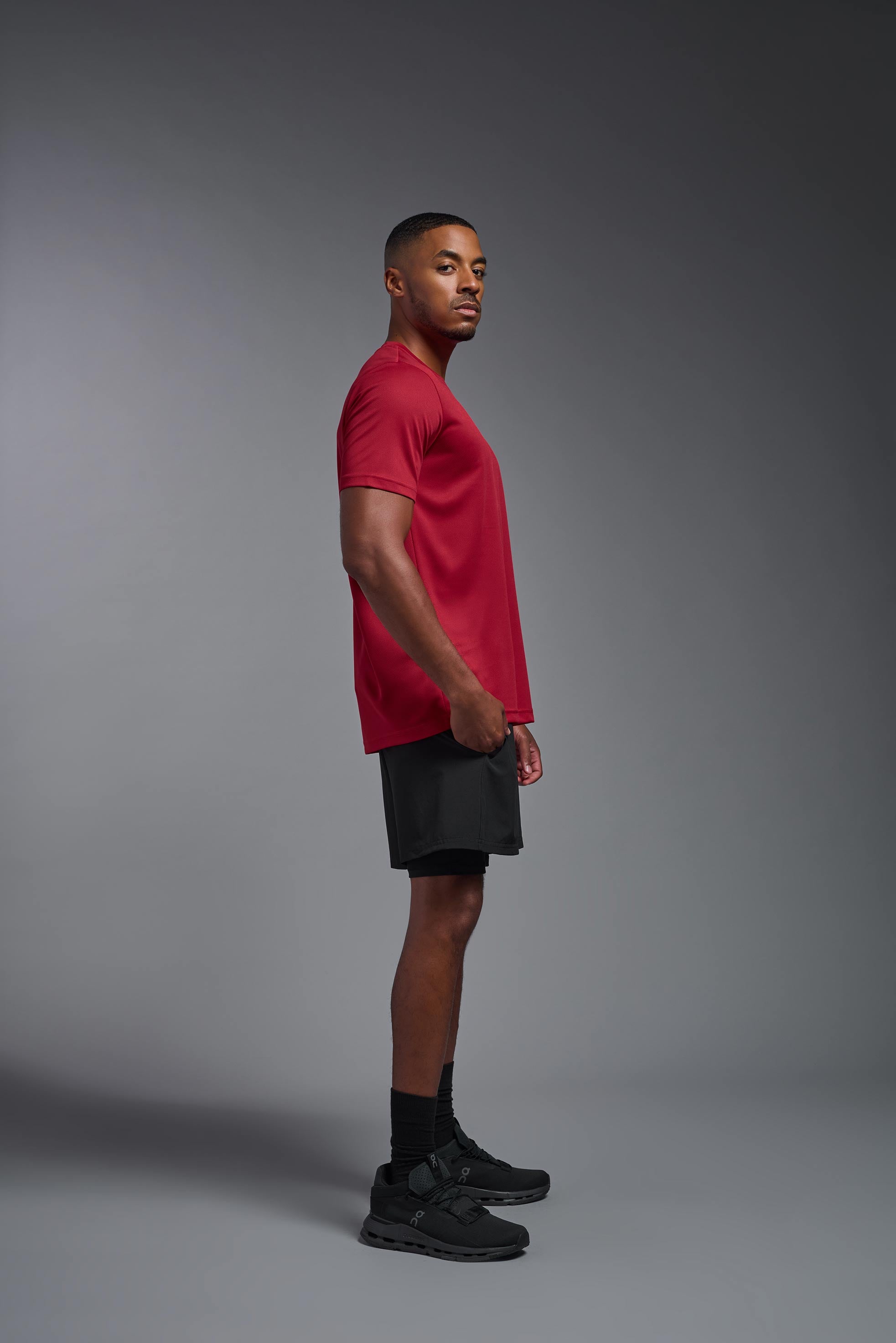 A male model wearing the men's Oxymesh Crewneck Tech Tee Imported Version in the color true red, made out of 100% polyester, showing the relaxed fit from a side view. This shirt is breathable, quick-drying, moisture-wicking, and Imported for a better price on bulk apparel. #true-red
