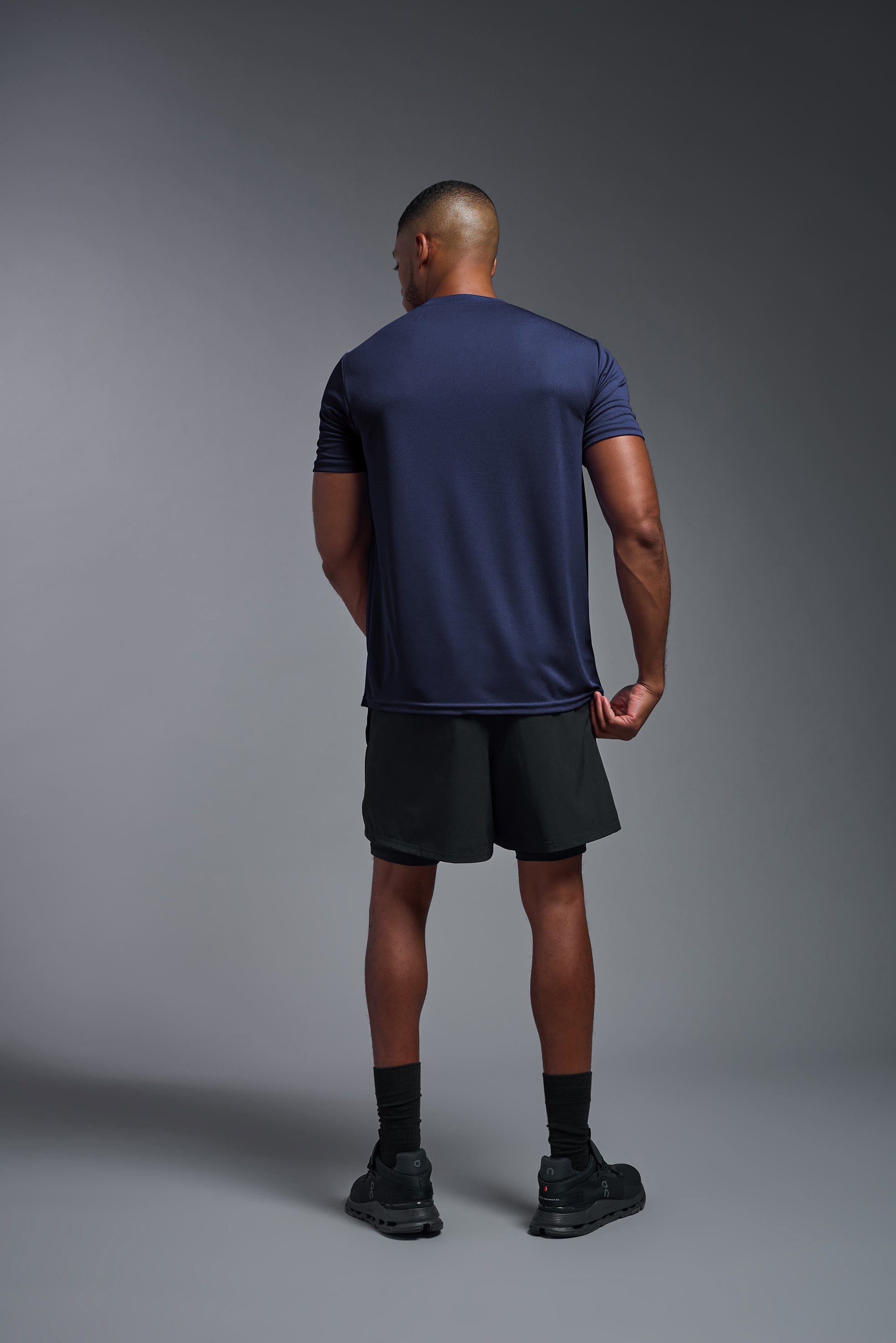 A male model wearing the men's Oxymesh Crewneck Tech Tee Imported Version in the color navy blue, made out of 100% polyester, showing the relaxed fit from a back view. This shirt is breathable, quick-drying, moisture-wicking, and Imported for a better price on bulk apparel. #navy-blue