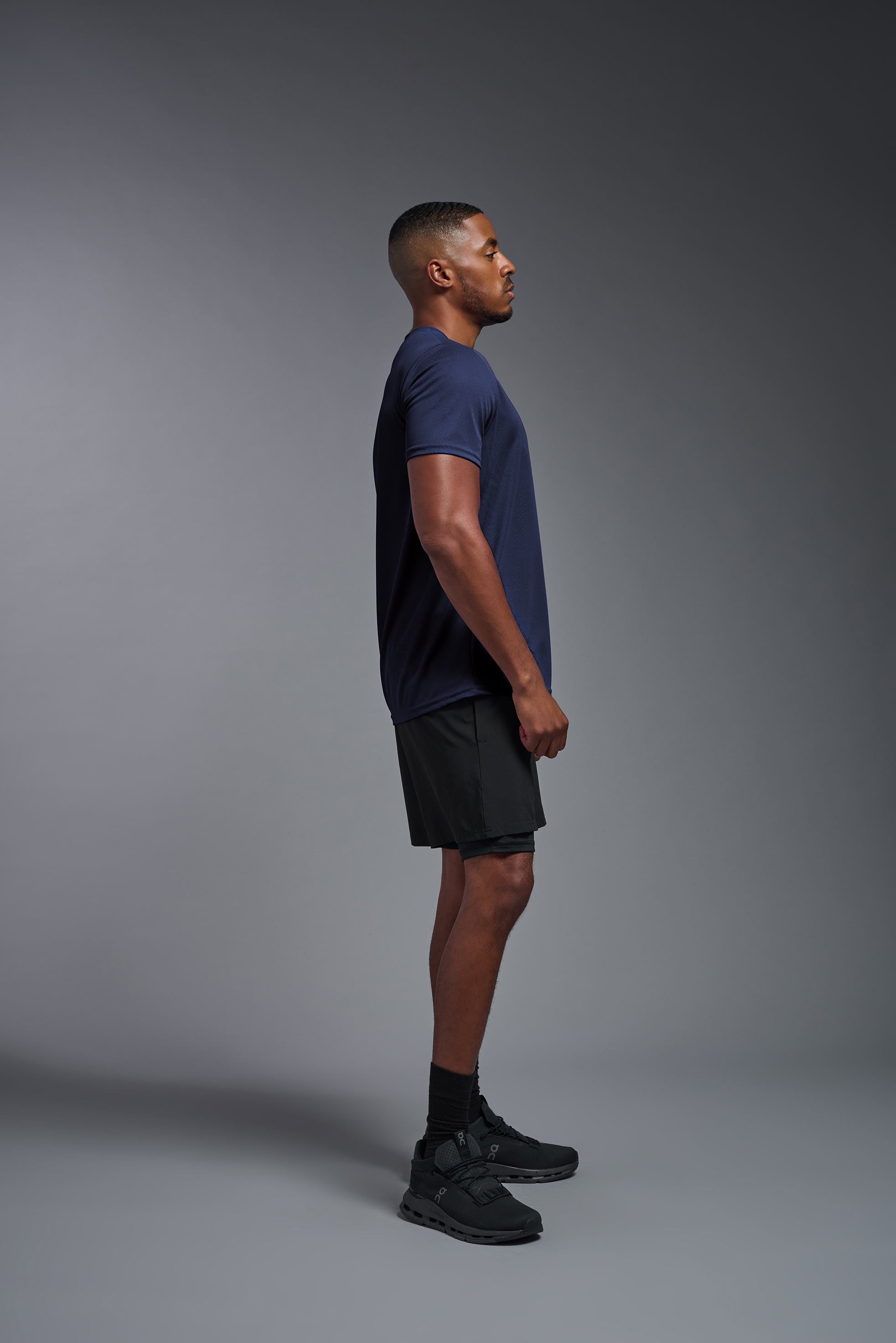 A male model wearing the men's Oxymesh Crewneck Tech Tee Imported Version in the color navy blue, made out of 100% polyester, showing the relaxed fit from a side view. This shirt is breathable, quick-drying, moisture-wicking, and Imported for a better price on bulk apparel. #navy-blue