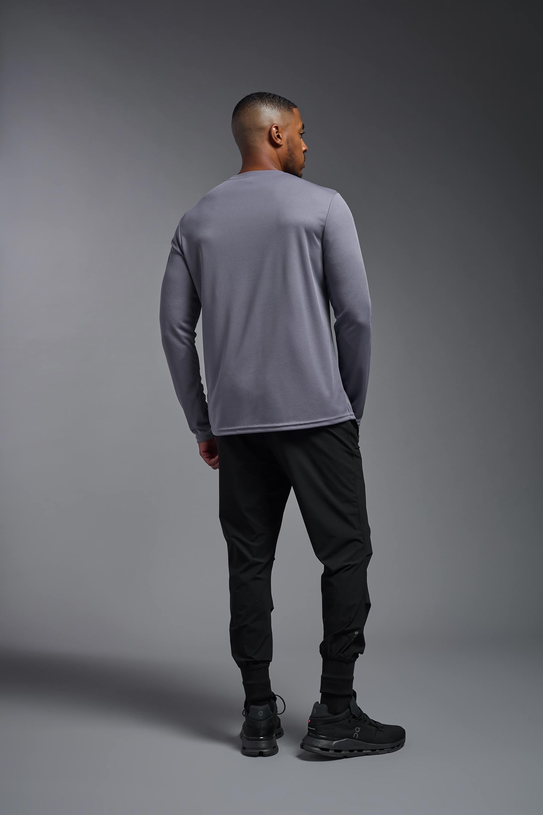 A male model wearing the men's Oxymesh Long Sleeve Tech Tee Imported Version in the color steel gray, made out of 100% polyester, showing the relaxed fit from a back view. This shirt is breathable, quick-drying, moisture-wicking, and Imported for a better price on bulk apparel. #steel-gray