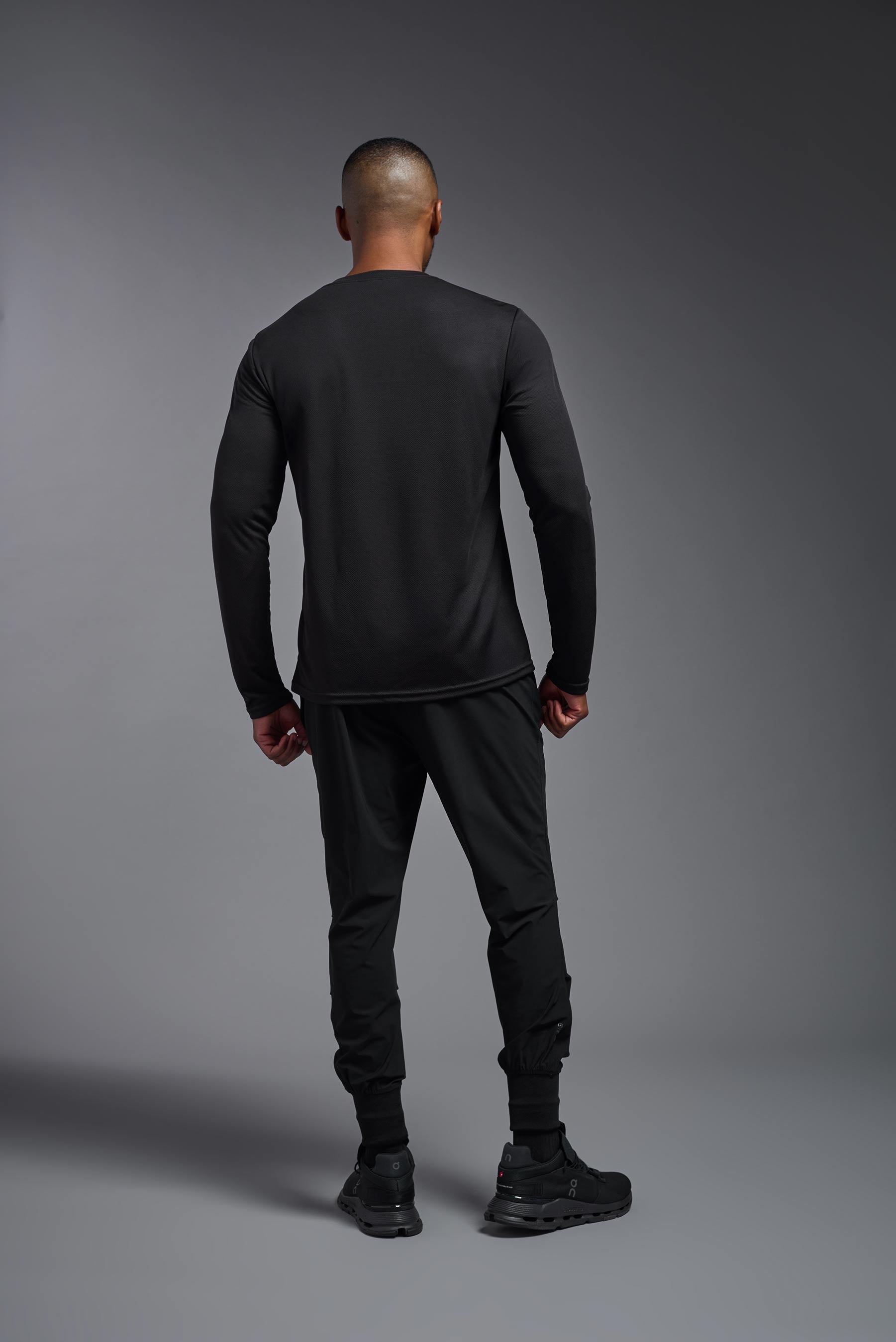 A male model wearing the men's Oxymesh Long Sleeve Tech Tee in the color black, made out of 100% polyester, showing the relaxed fit from a back view. This shirt is breathable, quick-drying, moisture-wicking, and made in the USA. #black