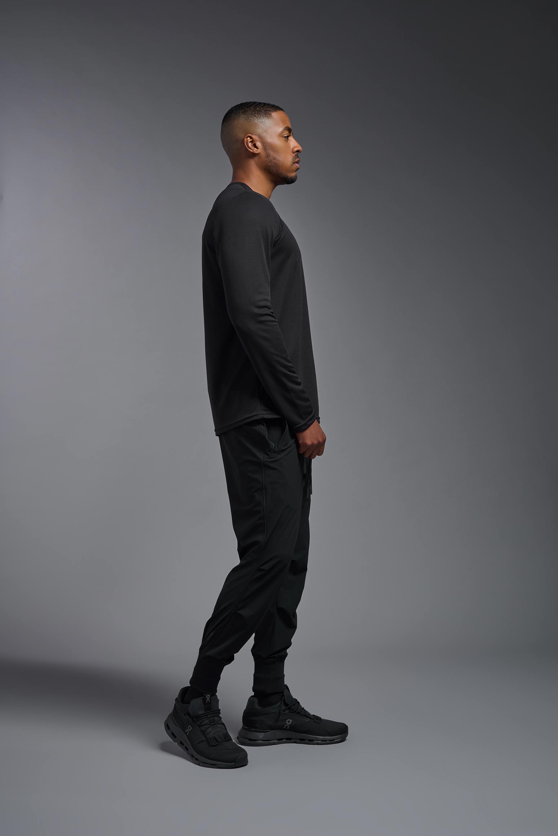 A male model wearing the men's Oxymesh Long Sleeve Tech Tee in the color black, made out of 100% polyester, showing the relaxed fit from a side view. This shirt is breathable, quick-drying, moisture-wicking, and made in the USA. #black