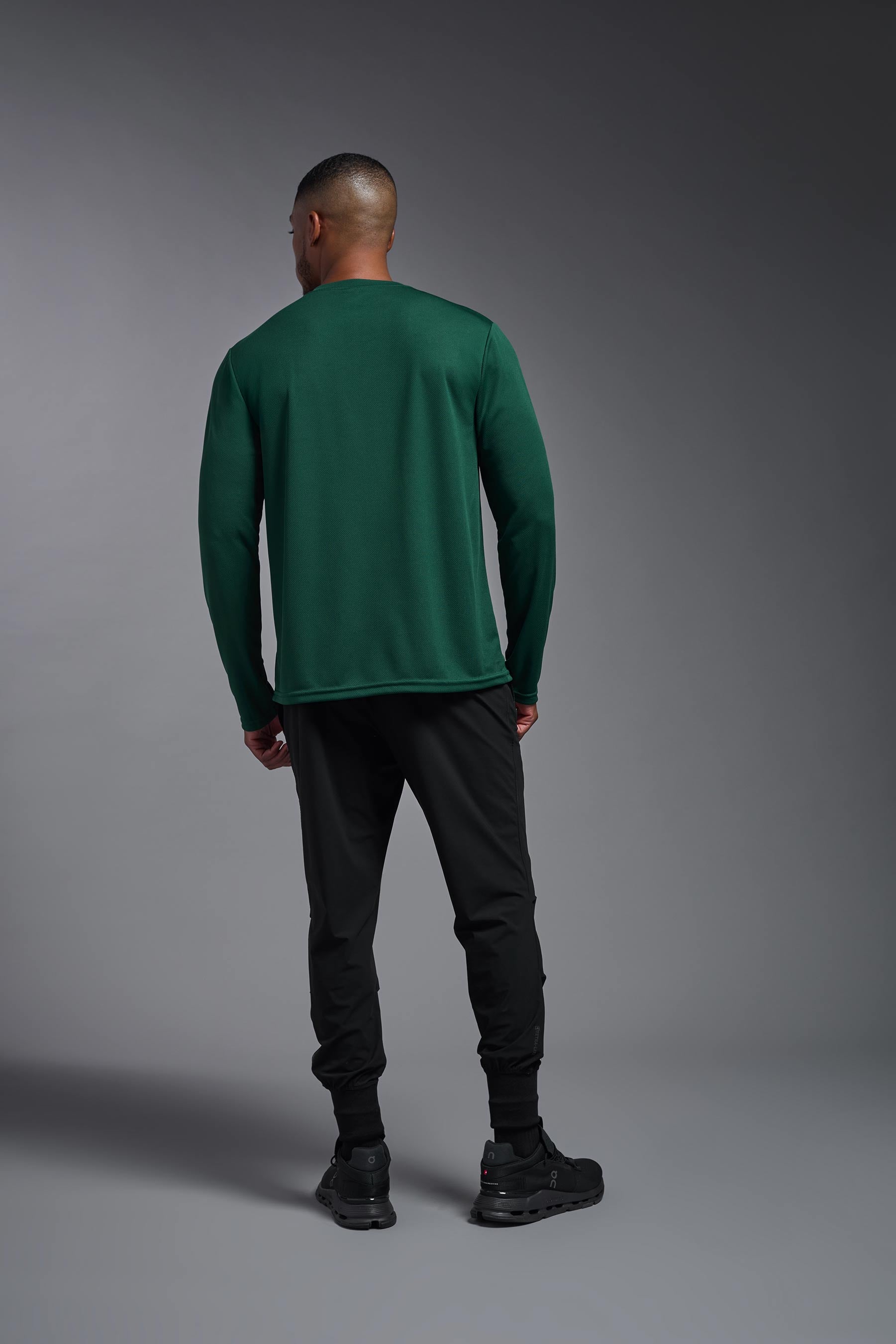 A male model wearing the men's Oxymesh Long Sleeve Tech Tee in the color forest green, made out of 100% polyester, showing the relaxed fit from a back view. This shirt is breathable, quick-drying, moisture-wicking, and made in the USA. #forest-green